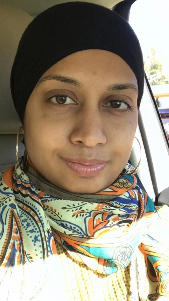 A woman wearing a black headscarf, large hoop earrings, and a colorful patterned scarf, sitting inside a vehicle with a window showing trees and a structure outside.