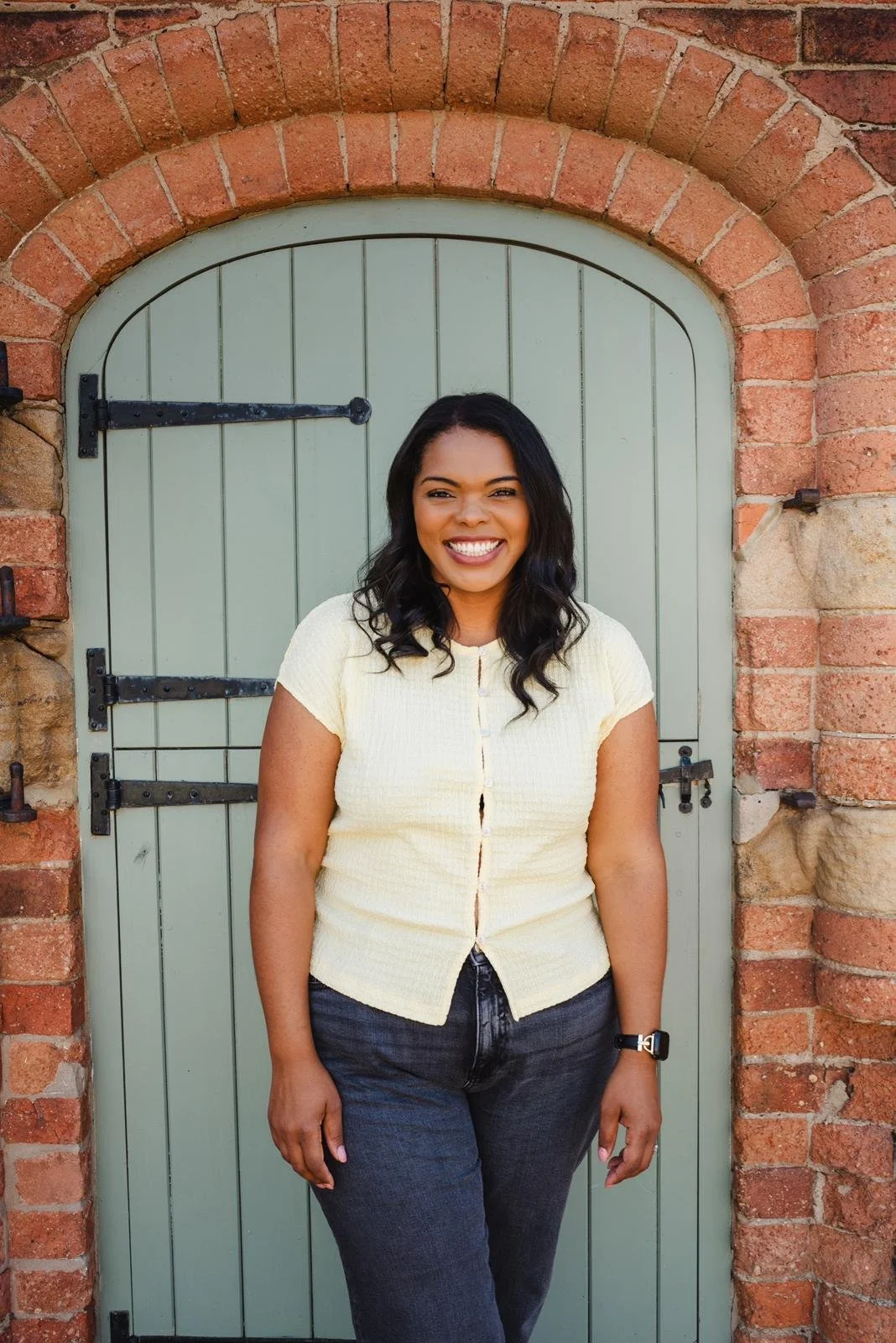 A woman with black hair smiling and standing in front of a light green arched door with black hardware, brick wall surrounding the door.