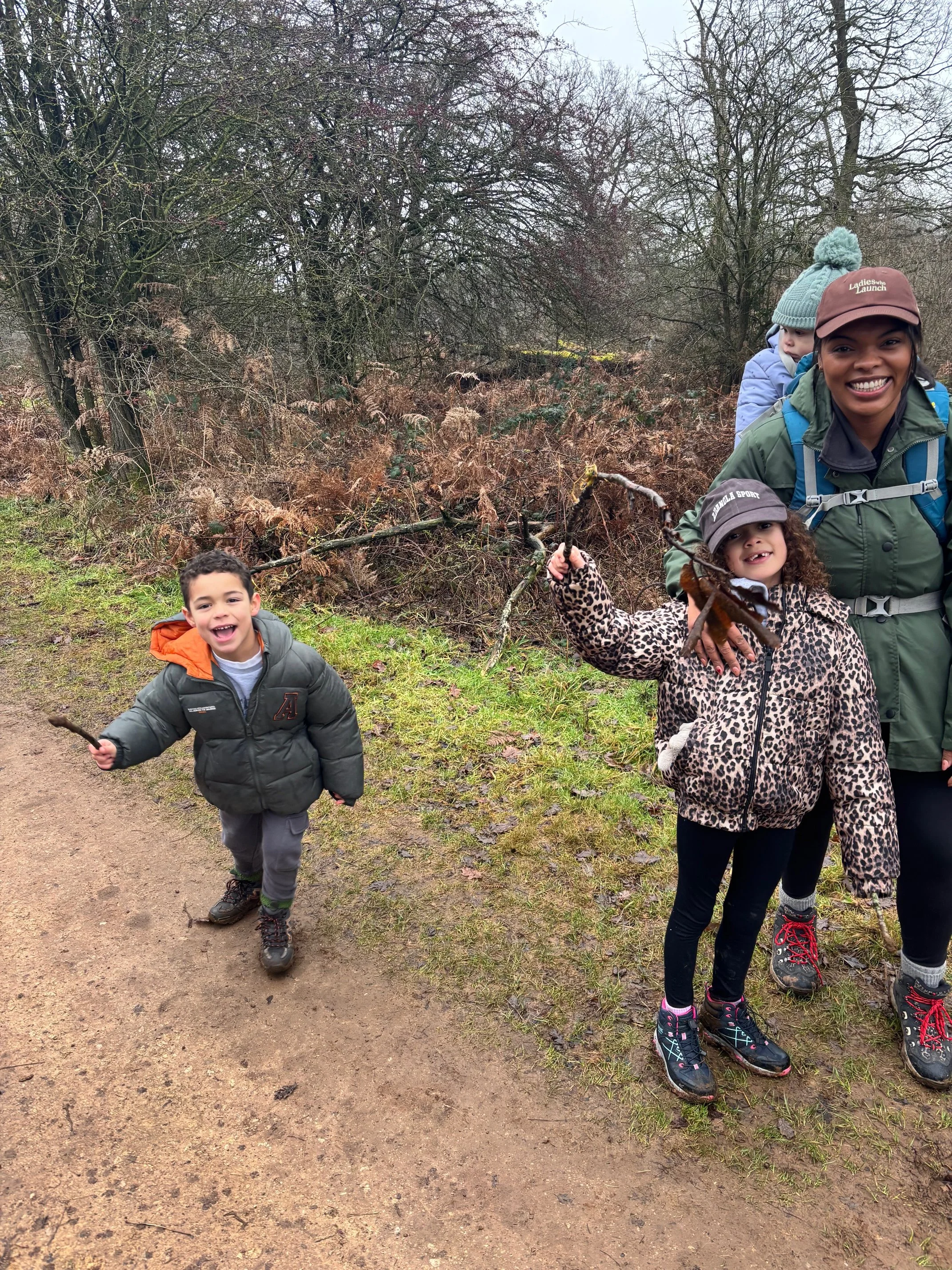 A group of children and a woman on a nature walk, the girl in leopard print holding a stick, others smiling, in a wooded area with bare trees and shrubs. A mother, busy founder, business woman and leader taking great care of her children.