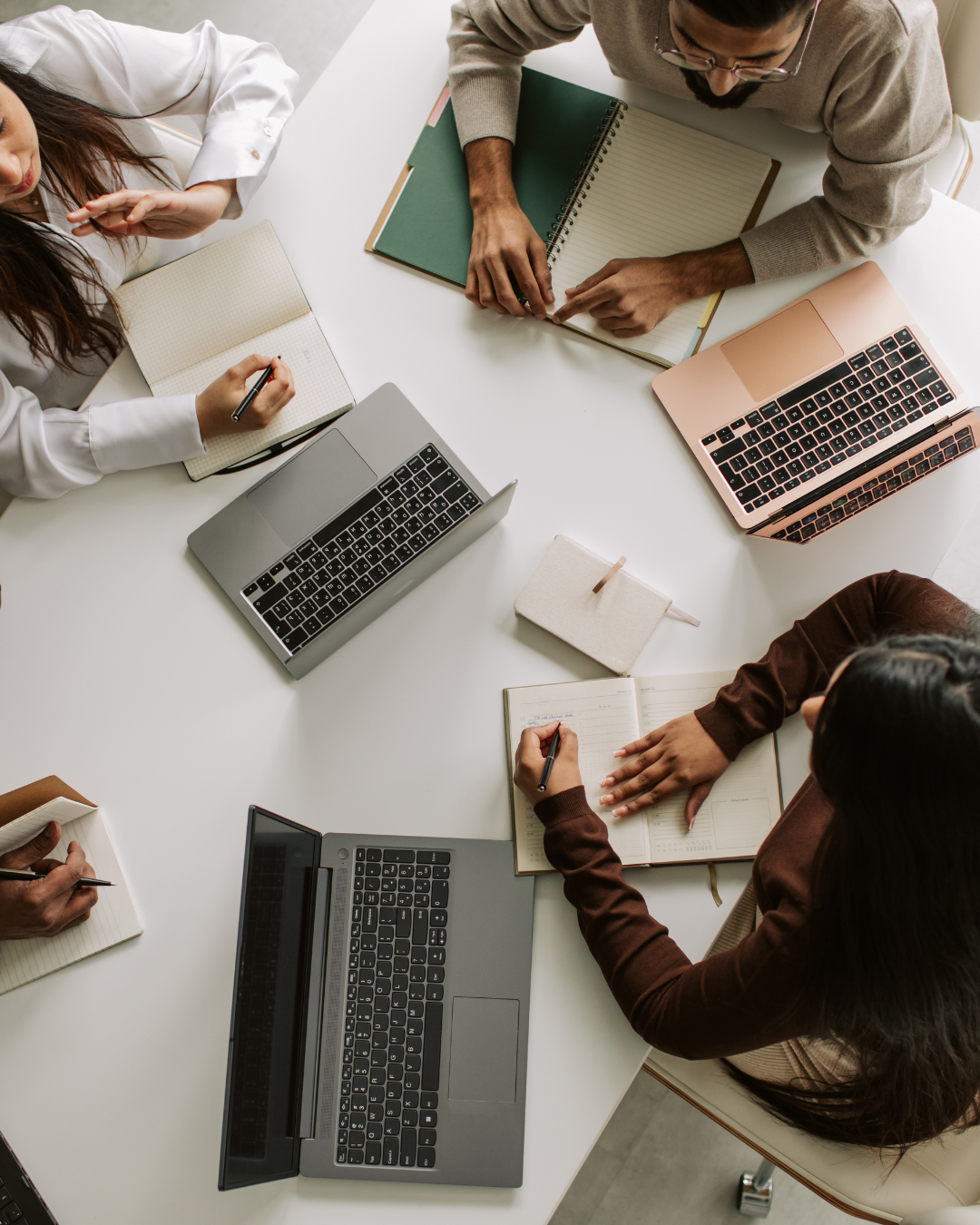 A top-down view of three people working at a white table with laptops, notebooks, and pens.