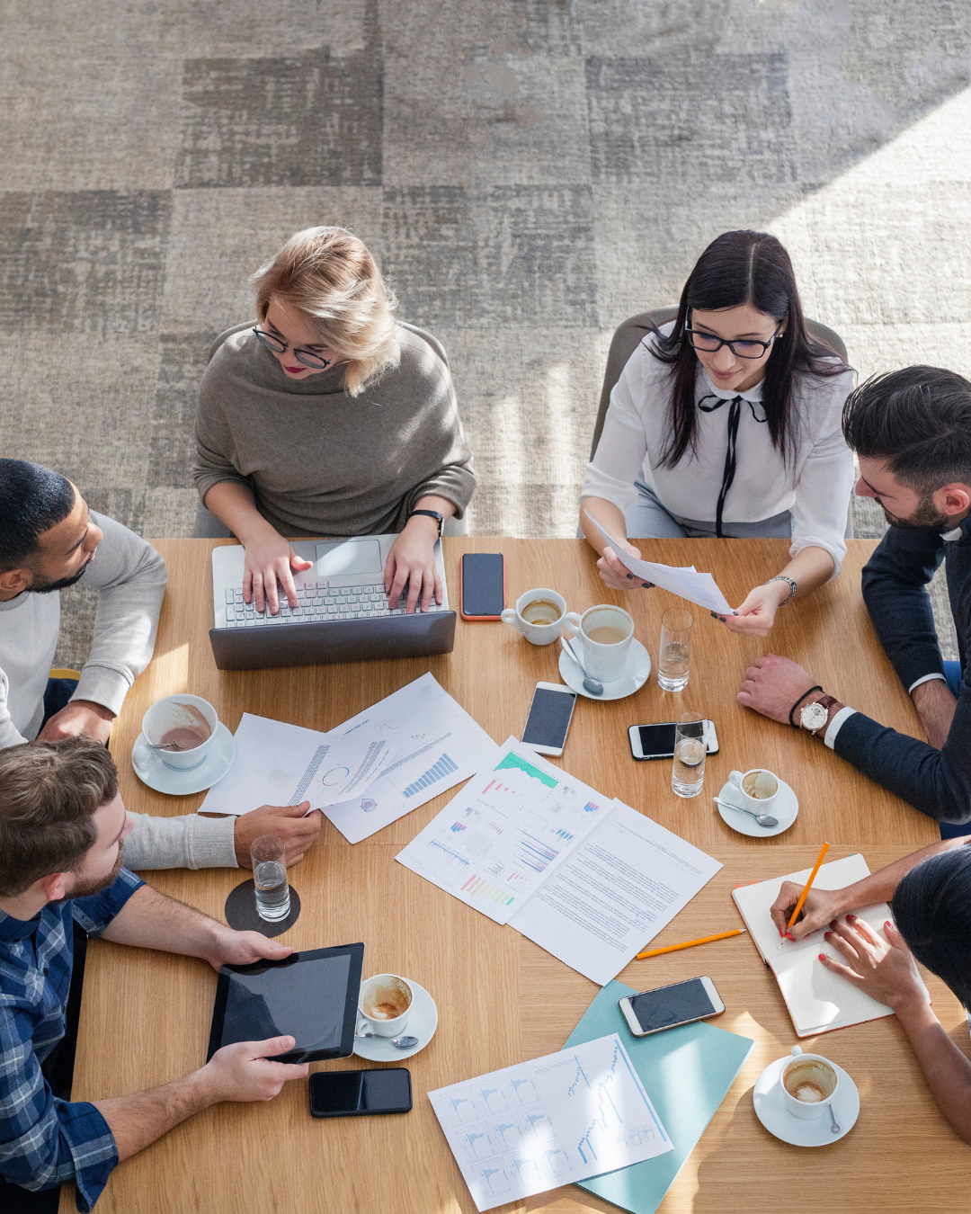 A group of six people having a meeting around a table with documents, cups of coffee, drinks, electronic devices, and writing materials.
