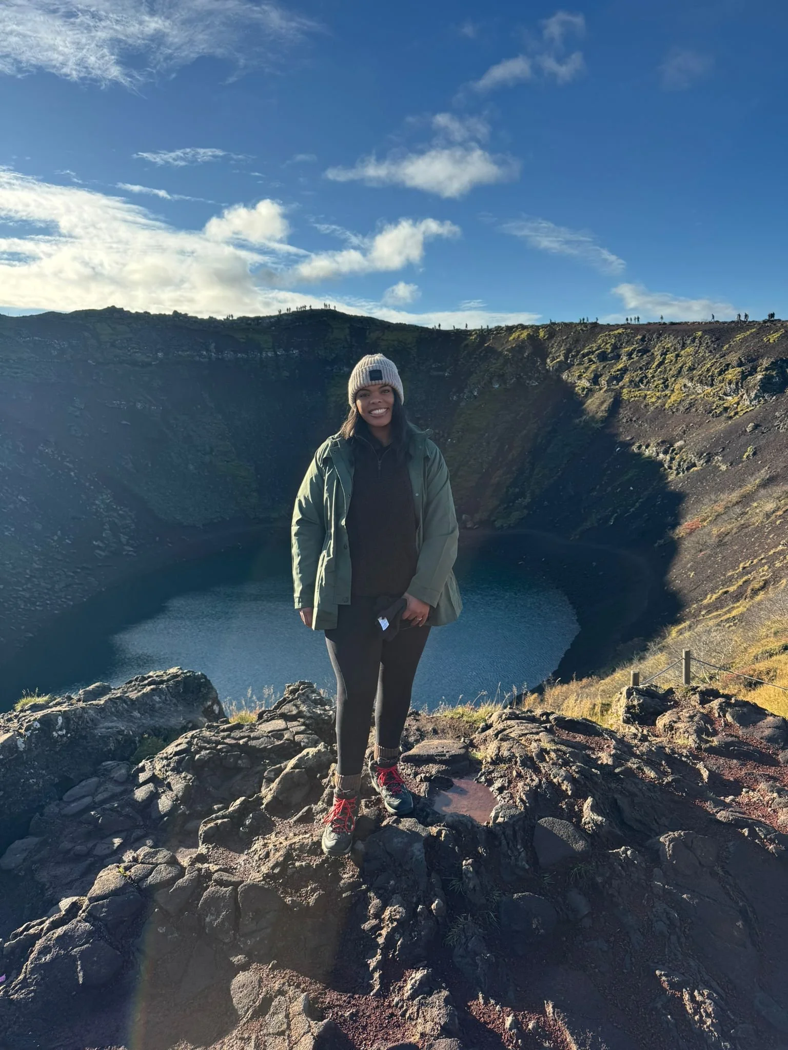 A woman in hiking gear standing on rocks in front of a crater lake with hillside and blue sky in the background. Leader, confidence, great mindset, calm and prepared.