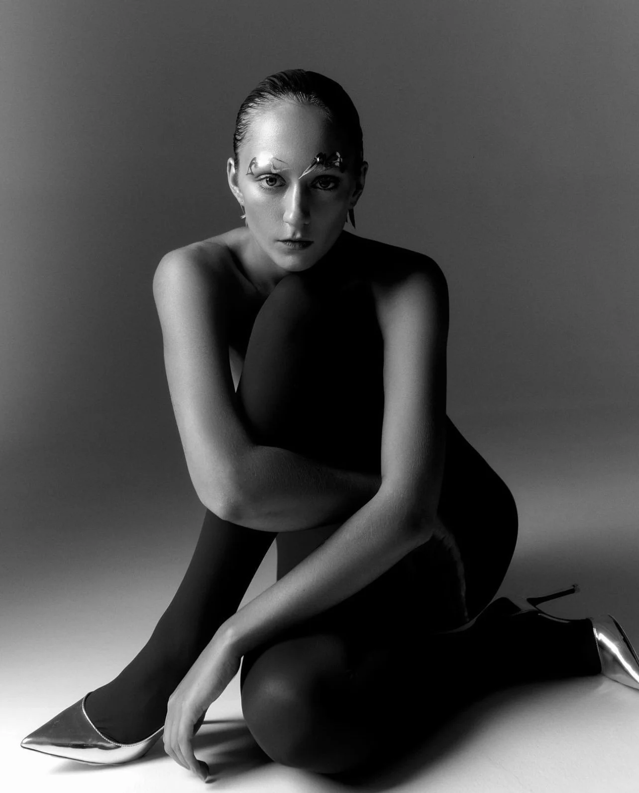 Black and white photo of a woman with slicked-back hair and dramatic makeup, sitting on the floor with one knee up and arms crossed, wearing shiny high-heeled shoes and a sleeveless top.
