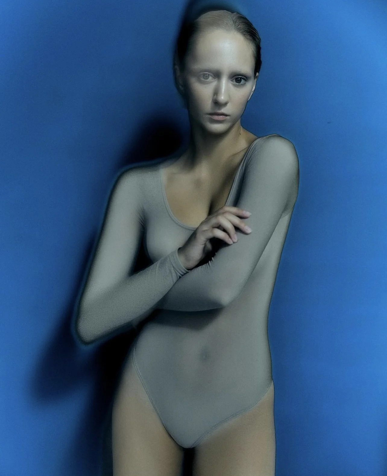 A woman with light skin and brown hair posing against a blue background, wearing a sheer taupe long-sleeve bodysuit.