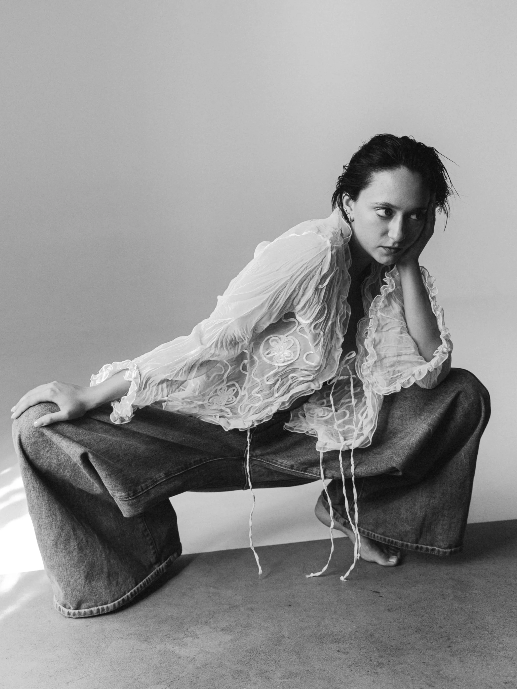 A woman with dark hair, wearing a sheer, embroidered blouse and wide-leg jeans, squatting against a plain wall, looking to the right with her head resting on her hand.