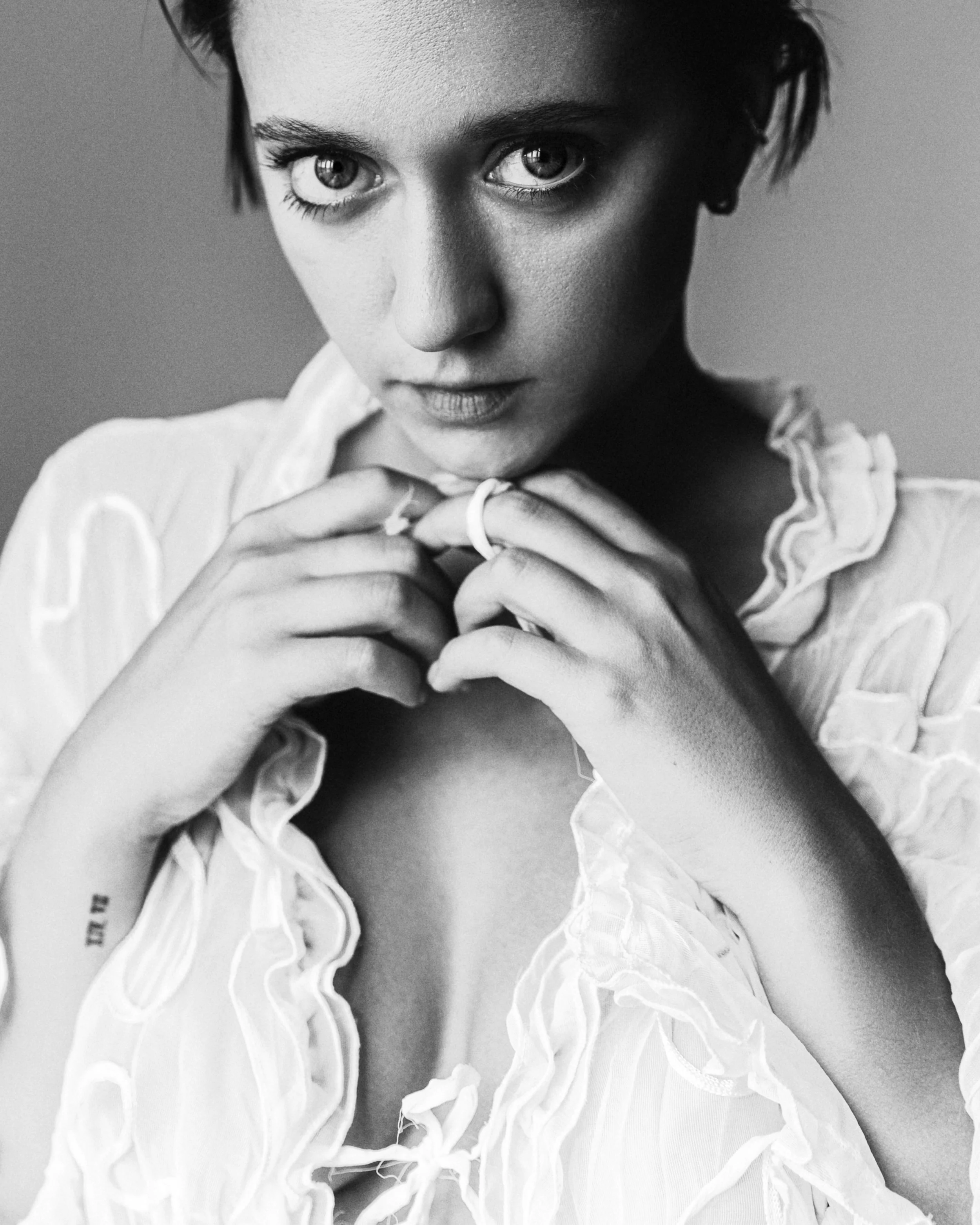 Black and white portrait of a woman with short hair looking directly at the camera, holding her hands near her collar, wearing a ruffled blouse.
