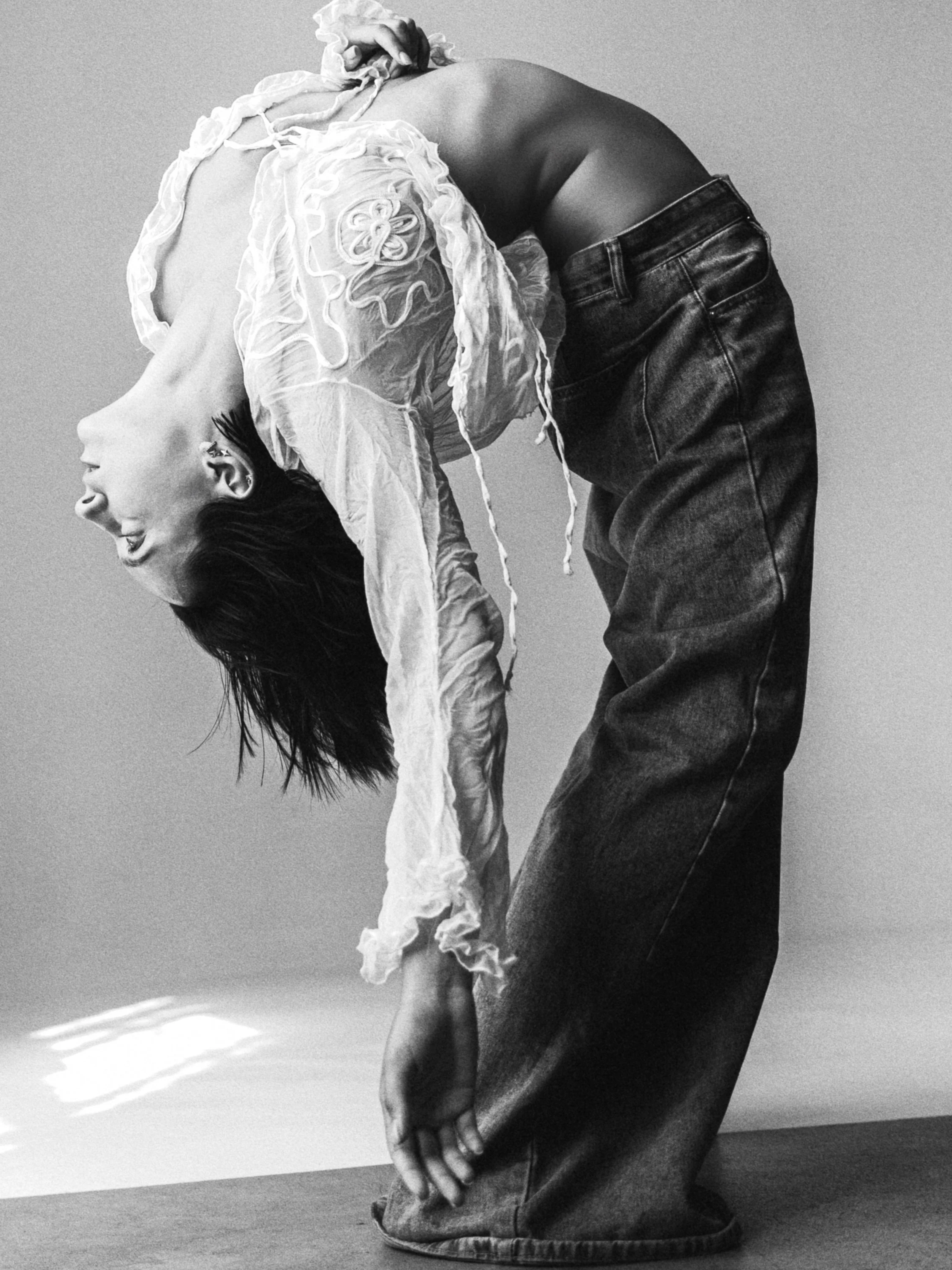 A person with dark hair is bending backwards, wearing a lace top and loose jeans, with their hair hanging down and one hand touching the ground.