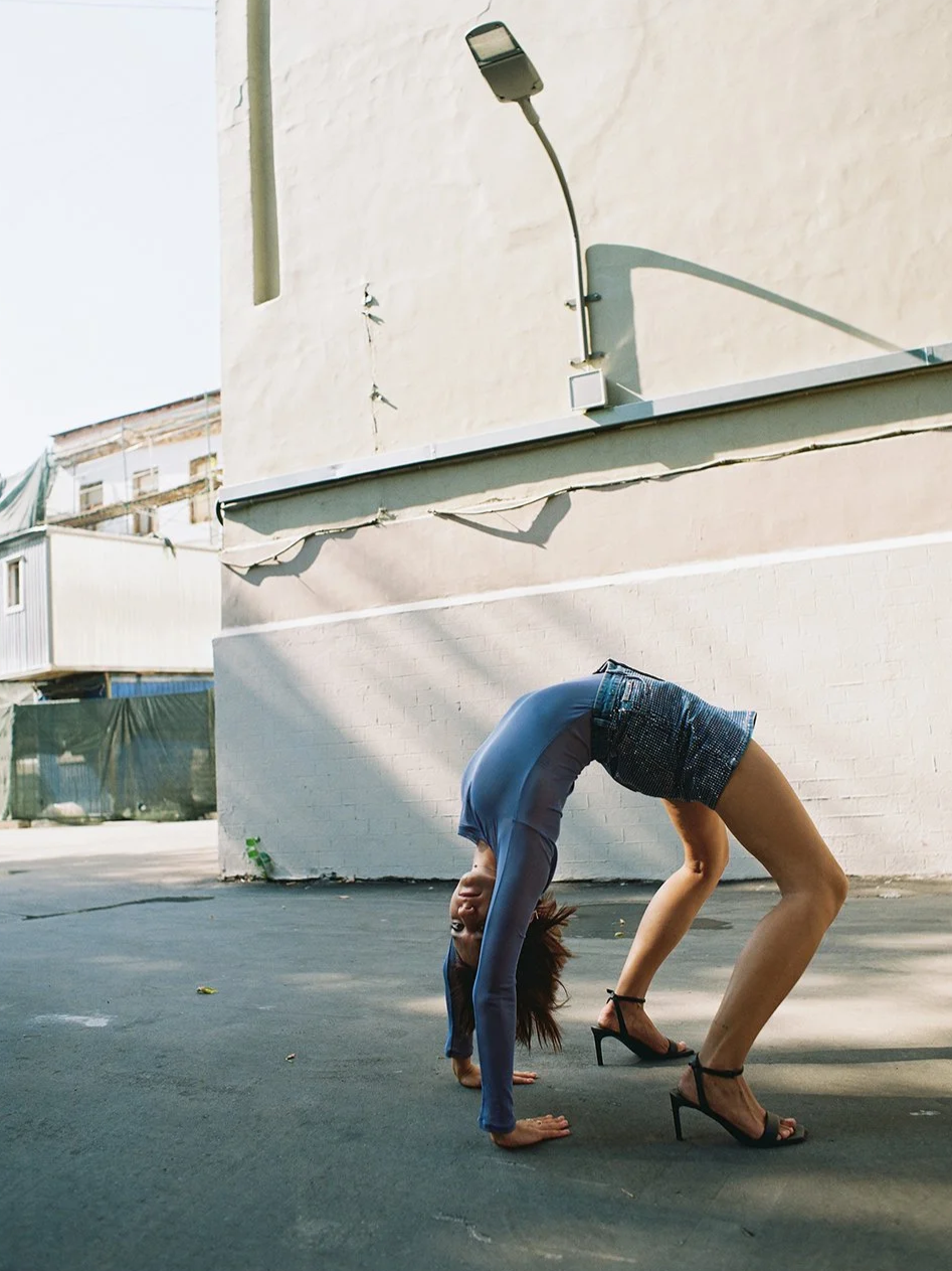 A woman in high heels bending backward in a bridge pose outdoors on a paved surface, with a white wall and building in the background.
