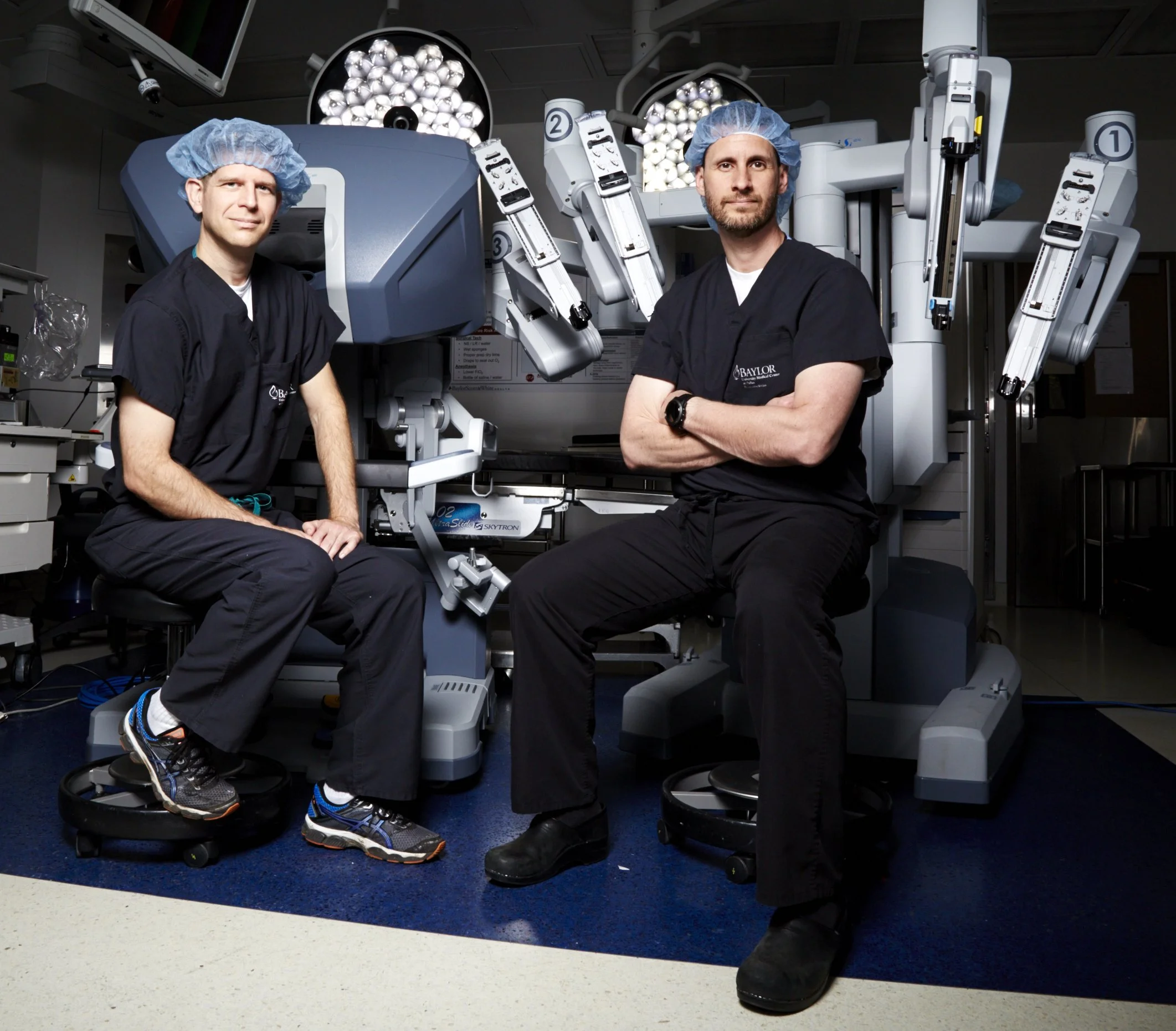 Two male surgeons in scrubs and blue surgical caps sitting in front of robotic surgical equipment in an operating room.
