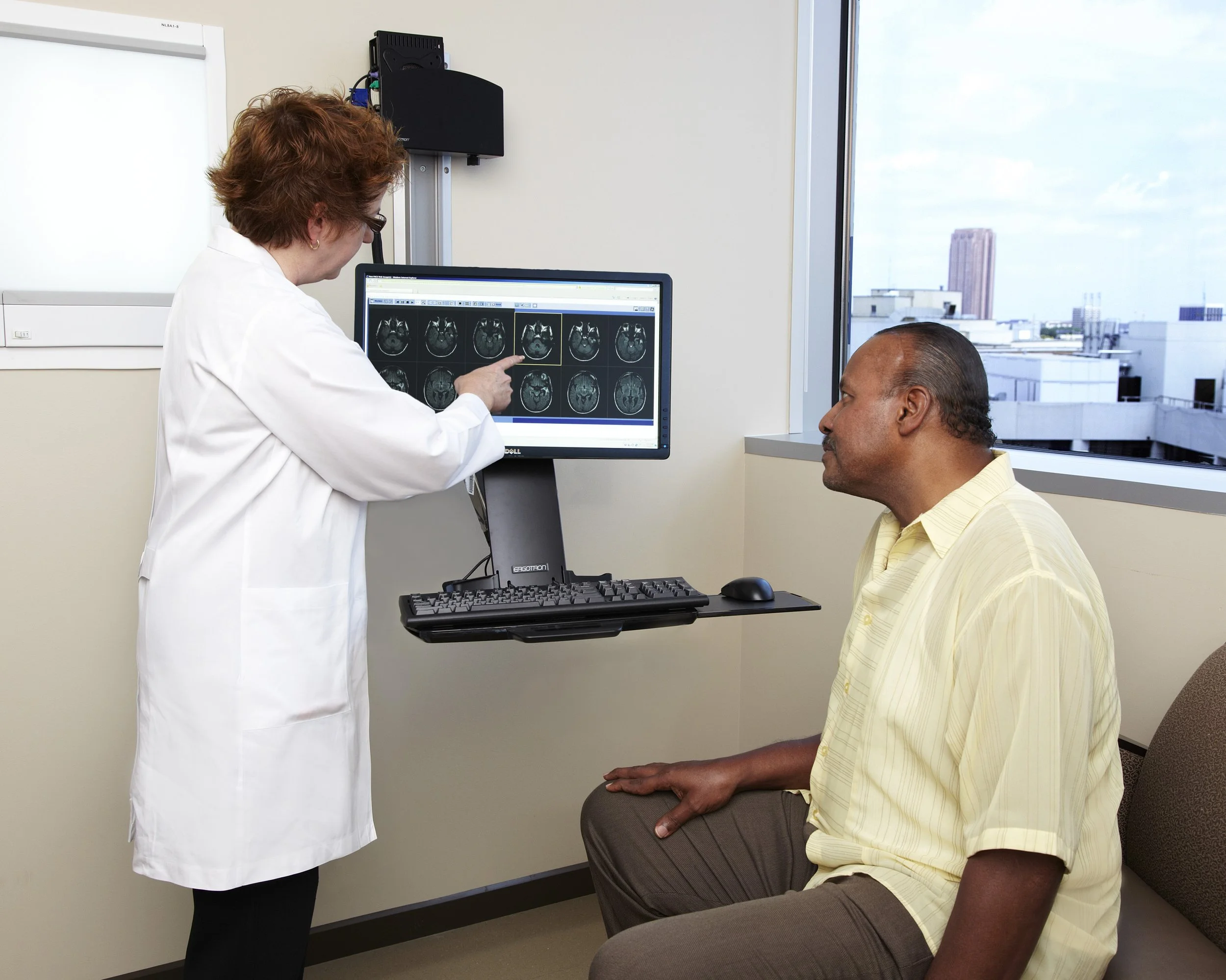 A doctor showing brain scan images on a computer monitor to a patient seated in a medical office with a large window.