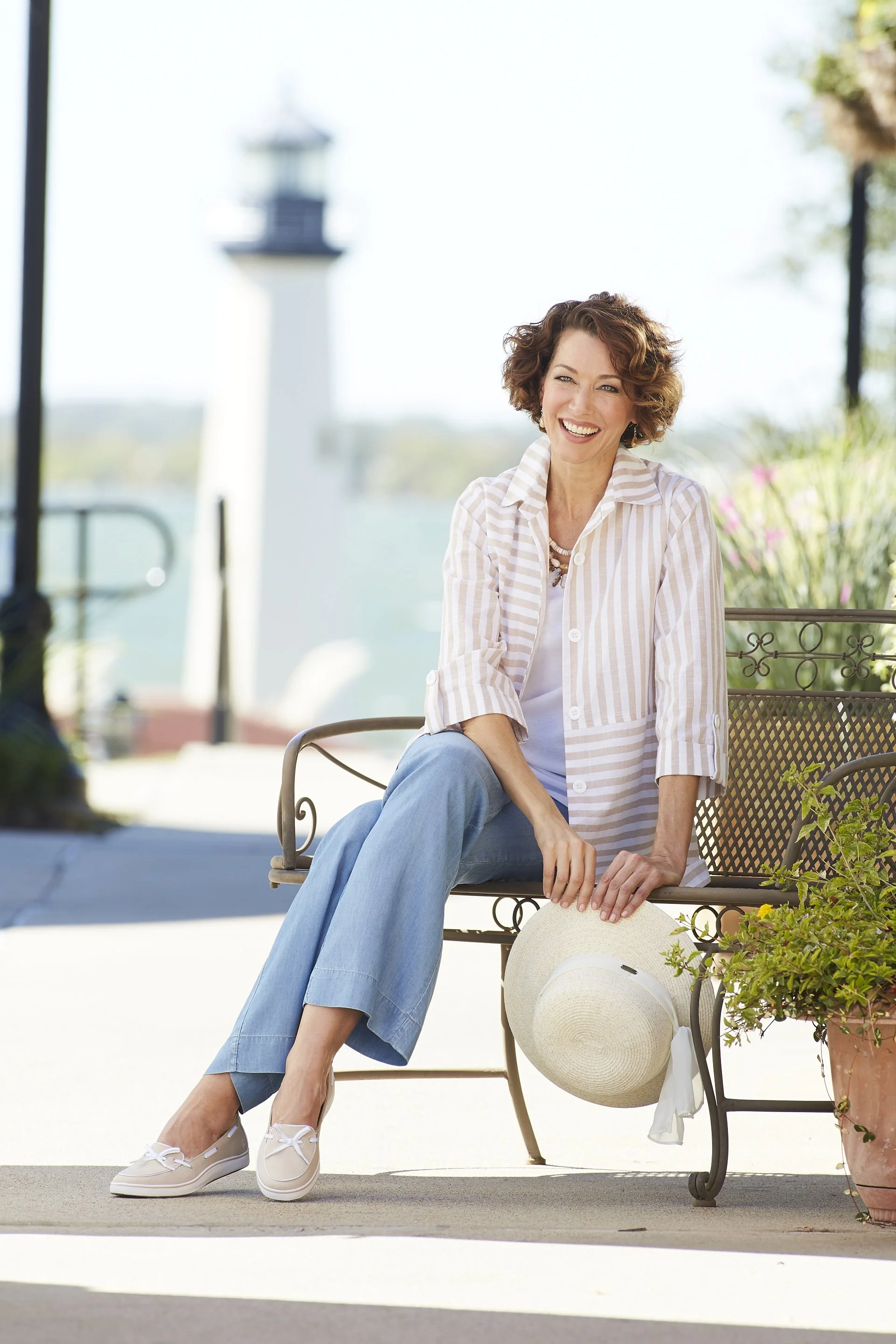 A smiling woman sitting on a metal bench outdoors, holding a wide-brimmed hat, with water and a lighthouse in the background.