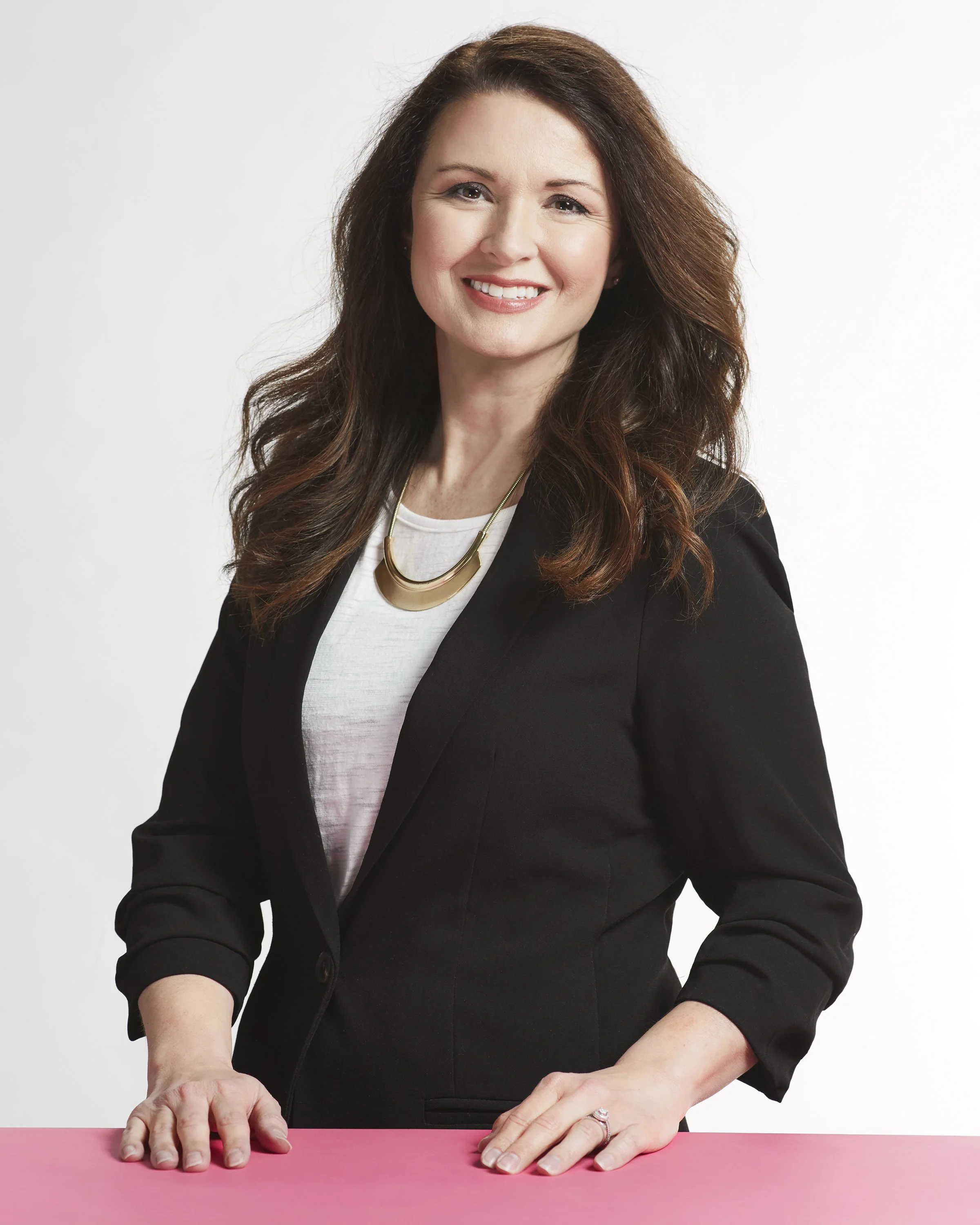 A woman with long, wavy brown hair, wearing a black blazer over a white top, smiling and leaning on a pink surface with her hands.