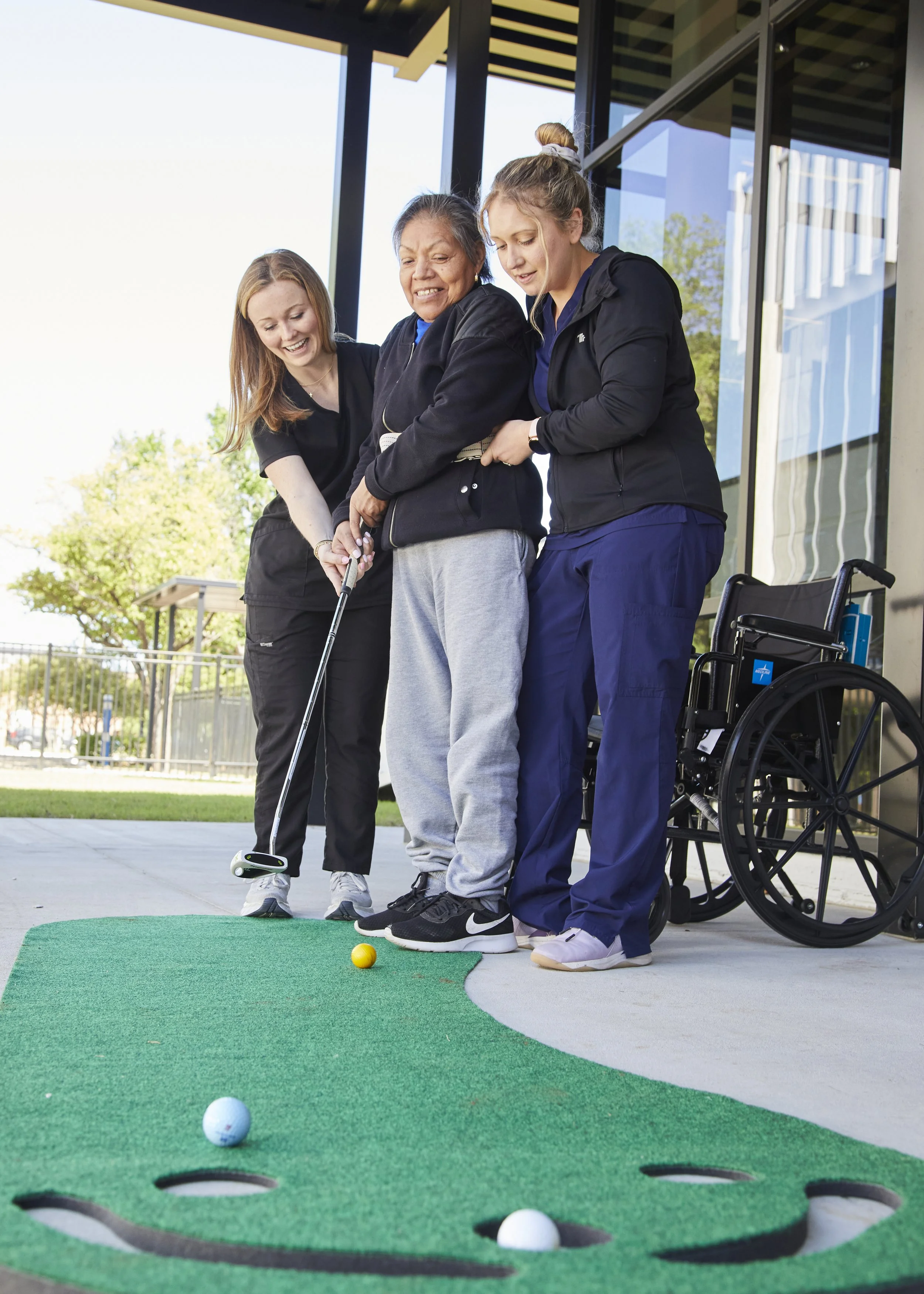 Three women, one in a wheelchair, enjoying a game of mini golf outdoors on a bright, sunny day.