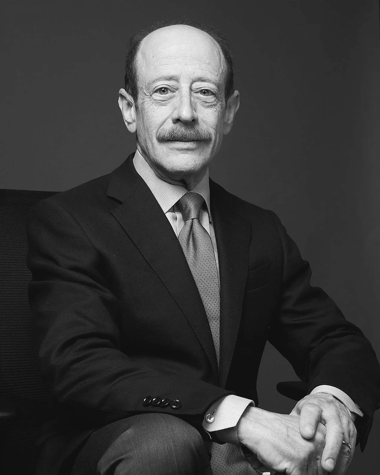 Black and white portrait of an older man with a mustache, wearing a suit and tie, seated with hands clasped.
