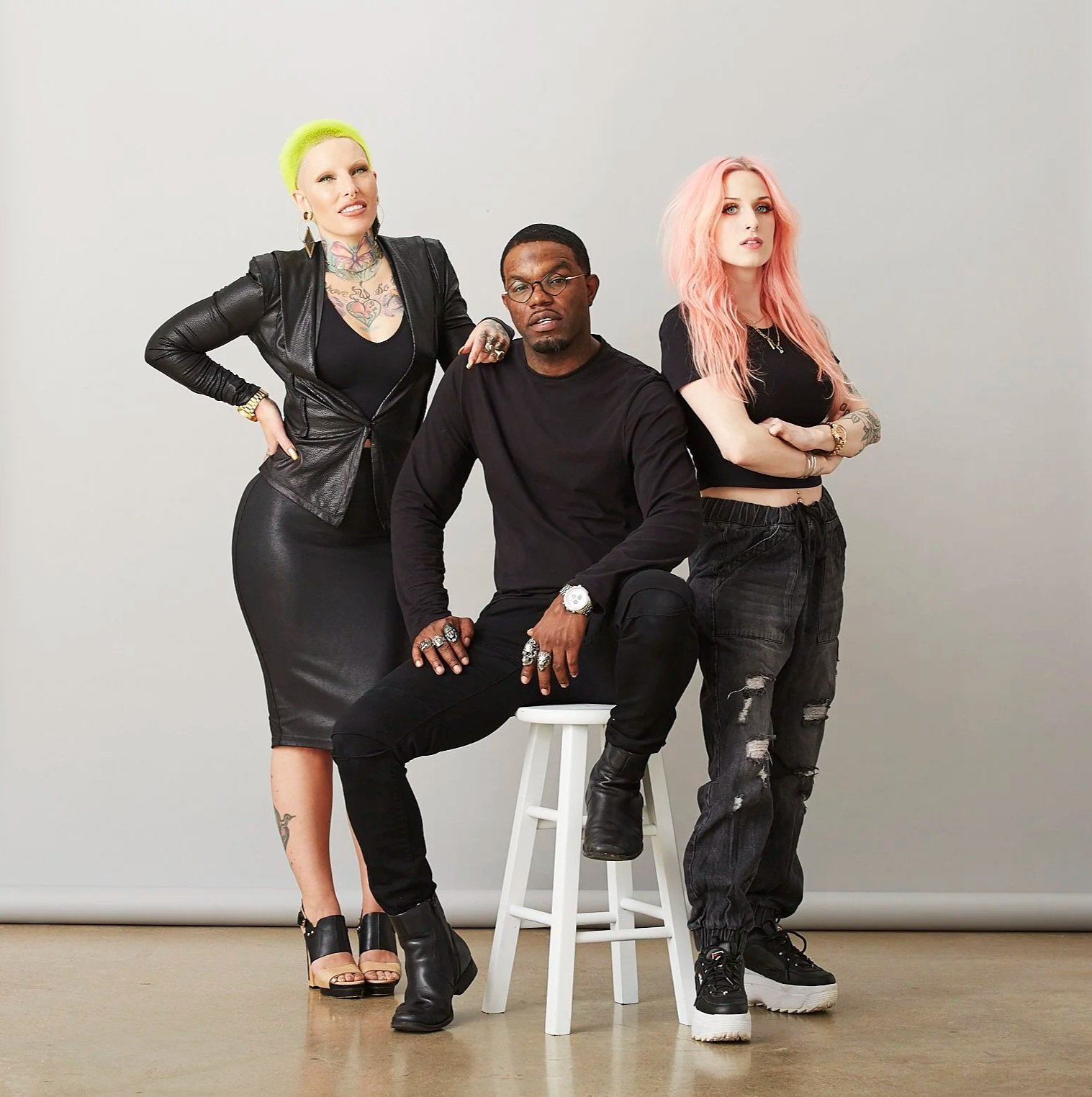 Three individuals posing together against a plain background. The woman on the left has bright neon green hair, tattoos, and is wearing a black leather jacket and a black skirt. The man in the center is sitting on a white stool, wears glasses, a blac
