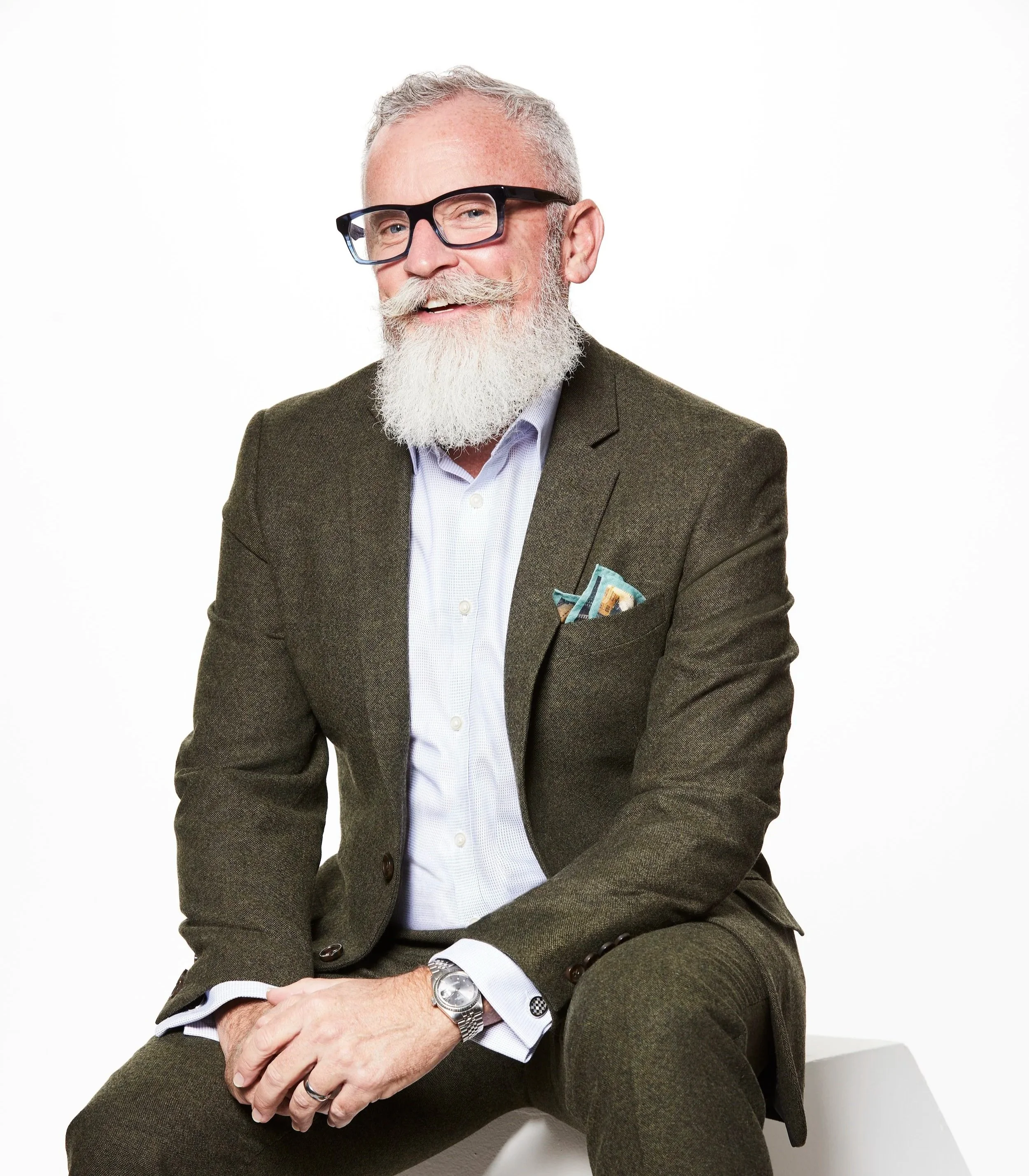 A smiling elderly man with gray hair, a beard, and glasses, wearing a dark green suit and a light blue shirt, sitting against a plain white background.