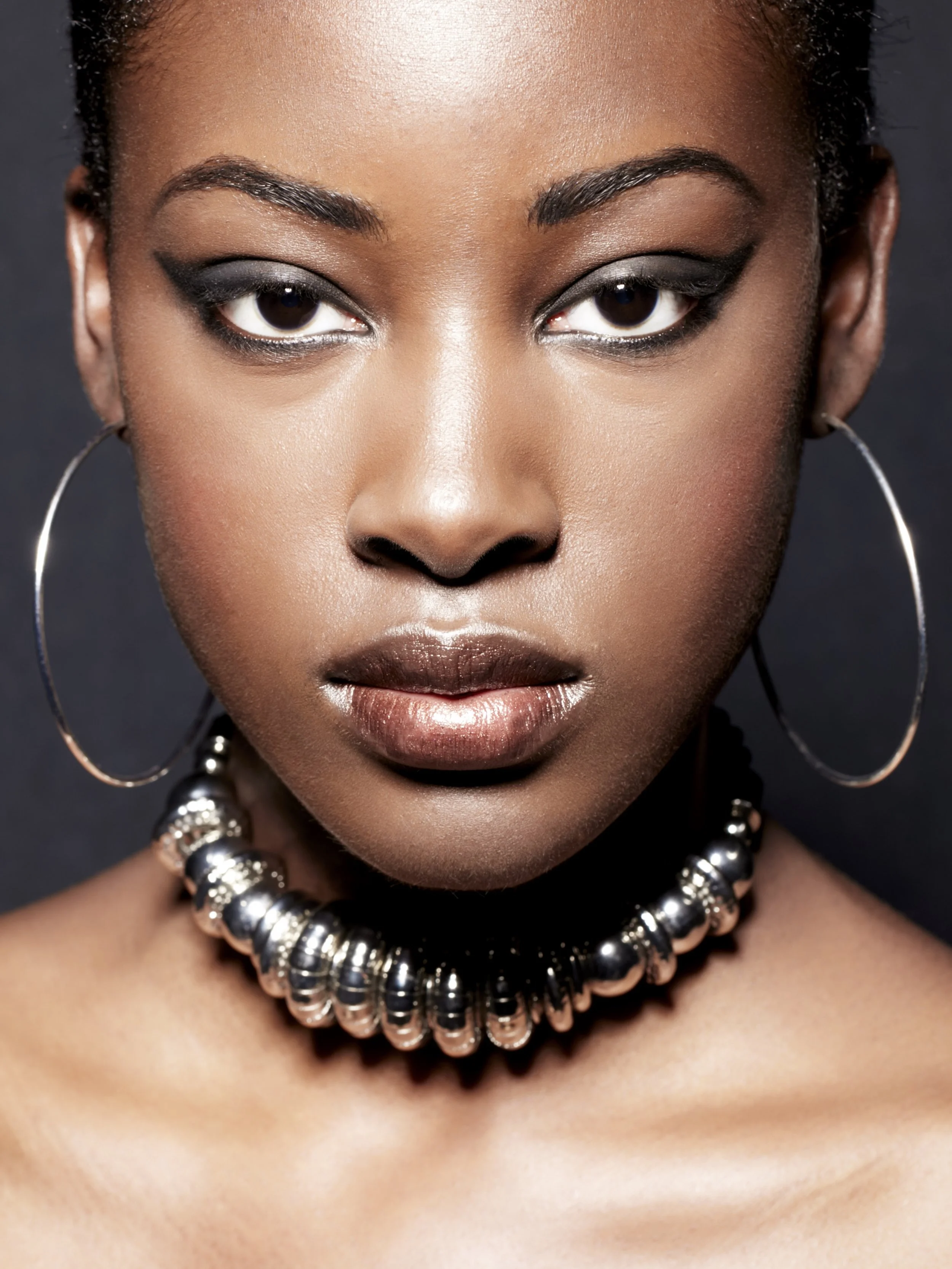 Close-up portrait of a woman with dark skin, wearing large hoop earrings and a metallic beaded choker necklace. She has makeup with dark eye shadow, eyeliner, and nude lipstick. Her hair is pulled back, and she is looking directly at the camera again