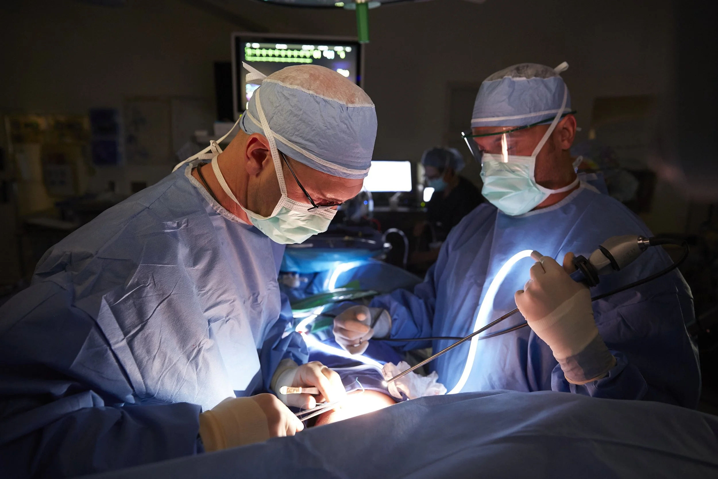 Surgeons in operating room performing surgery, wearing surgical gowns, masks, gloves, and protective eyewear, with bright lights and medical equipment around.
