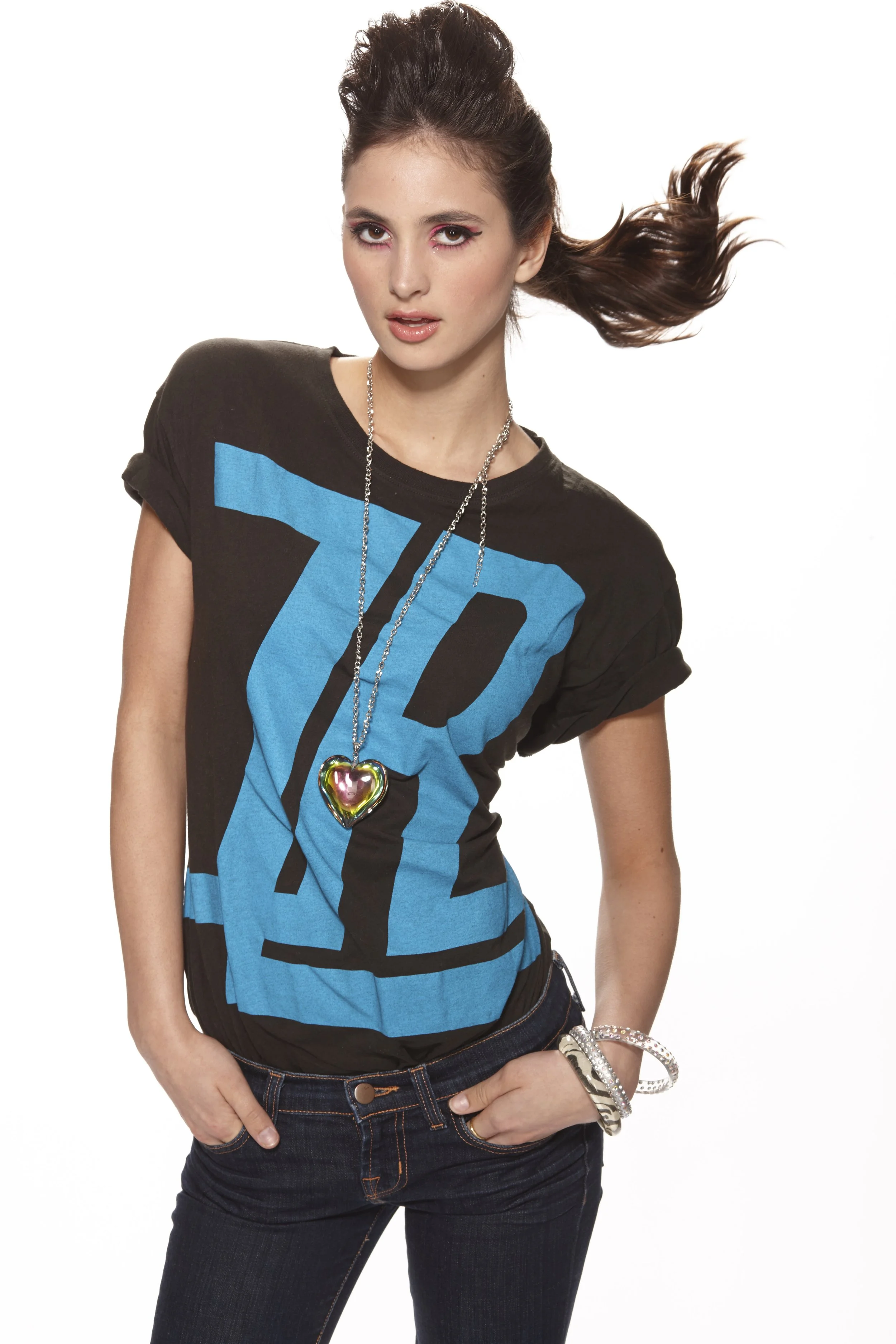 A young woman with long brown hair, styled with a voluminous waves and tied upward, is wearing a black T-shirt with a blue geometric design, dark jeans, and accessories including a long chain necklace with a heart-shaped pendant and several bracelets