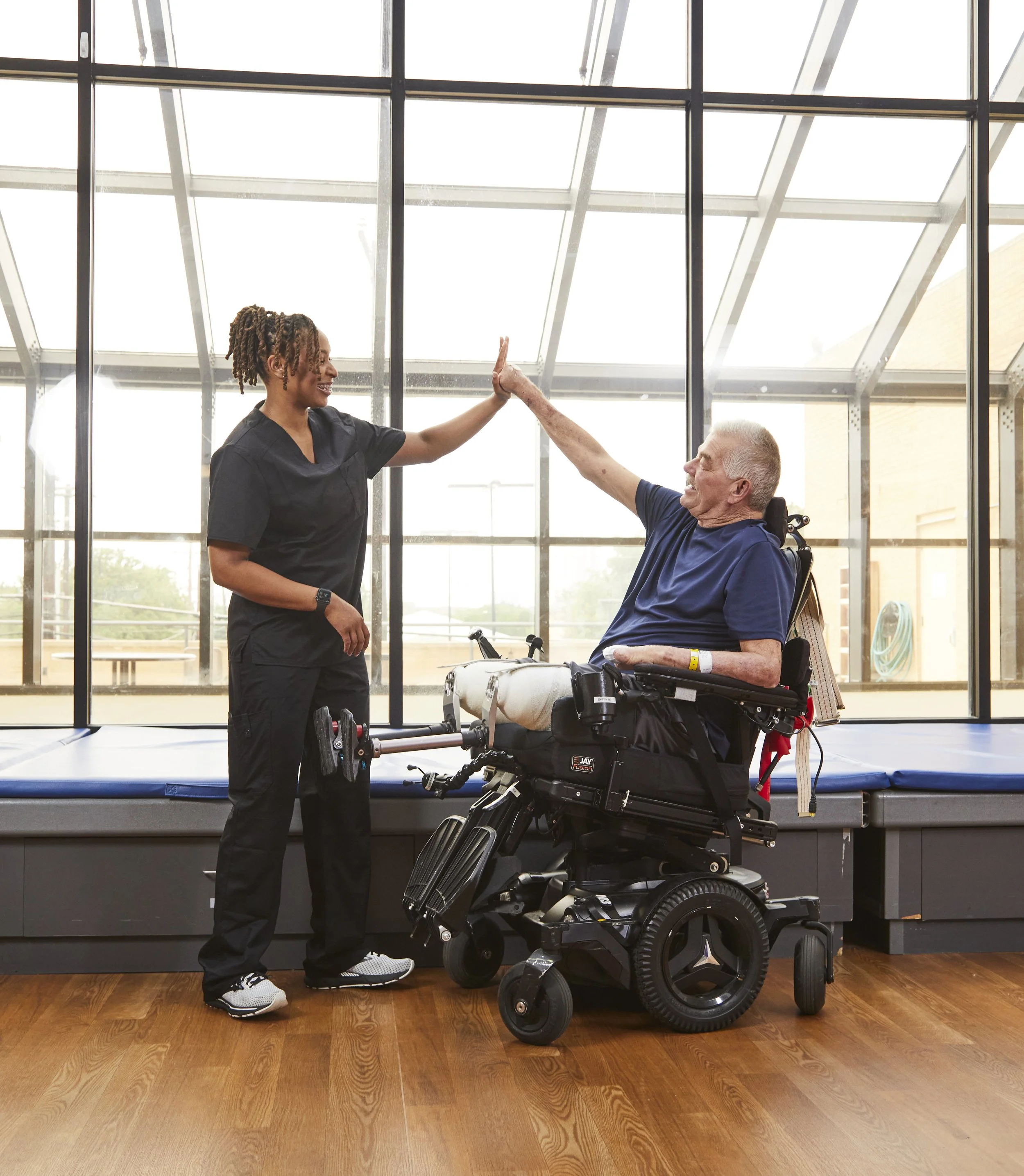 A healthcare worker giving a high five to an elderly man in a wheelchair inside a bright room with large glass windows.