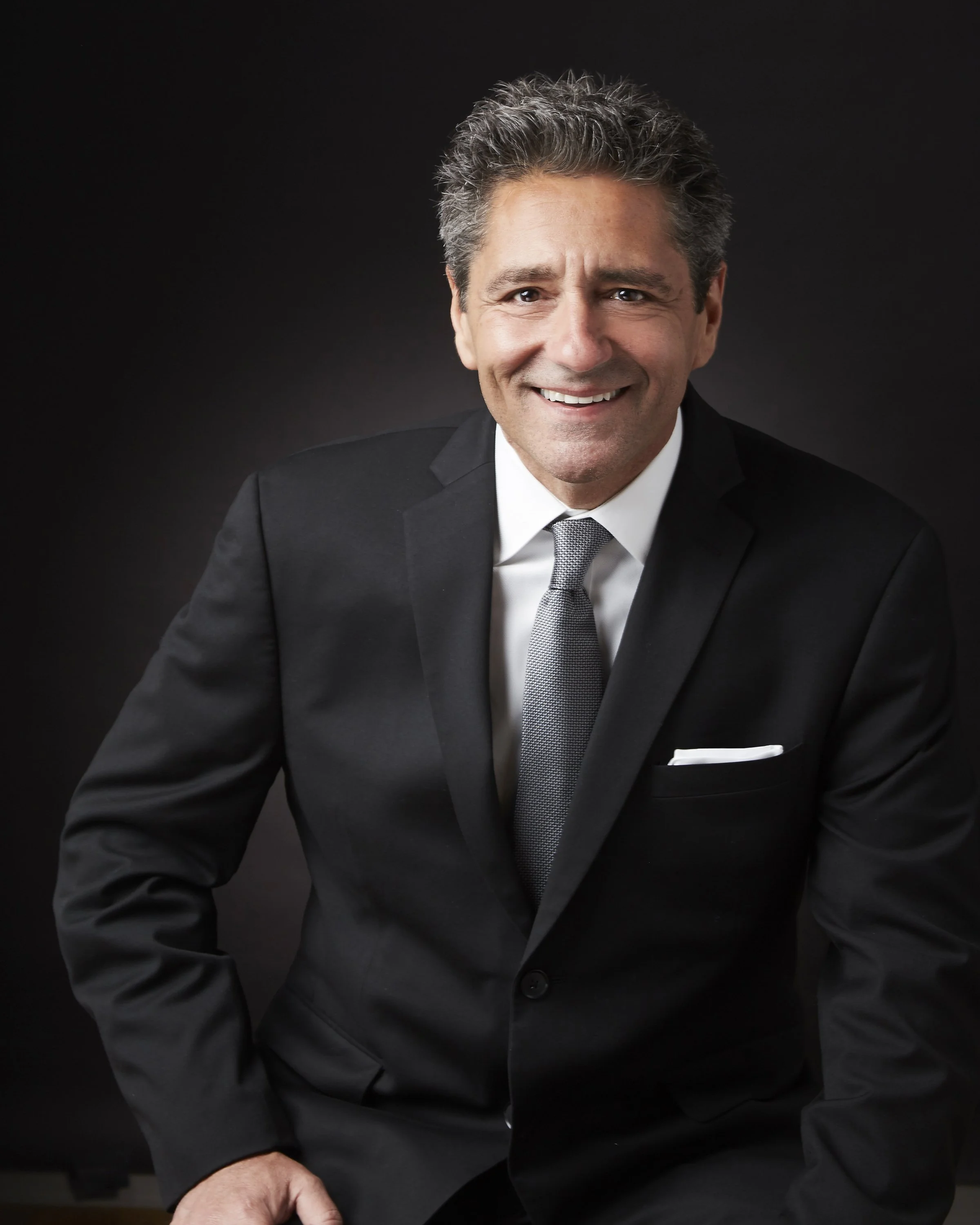 Portrait of a middle-aged man with gray hair, smiling, wearing a black suit, white shirt, and gray tie, against a black background.