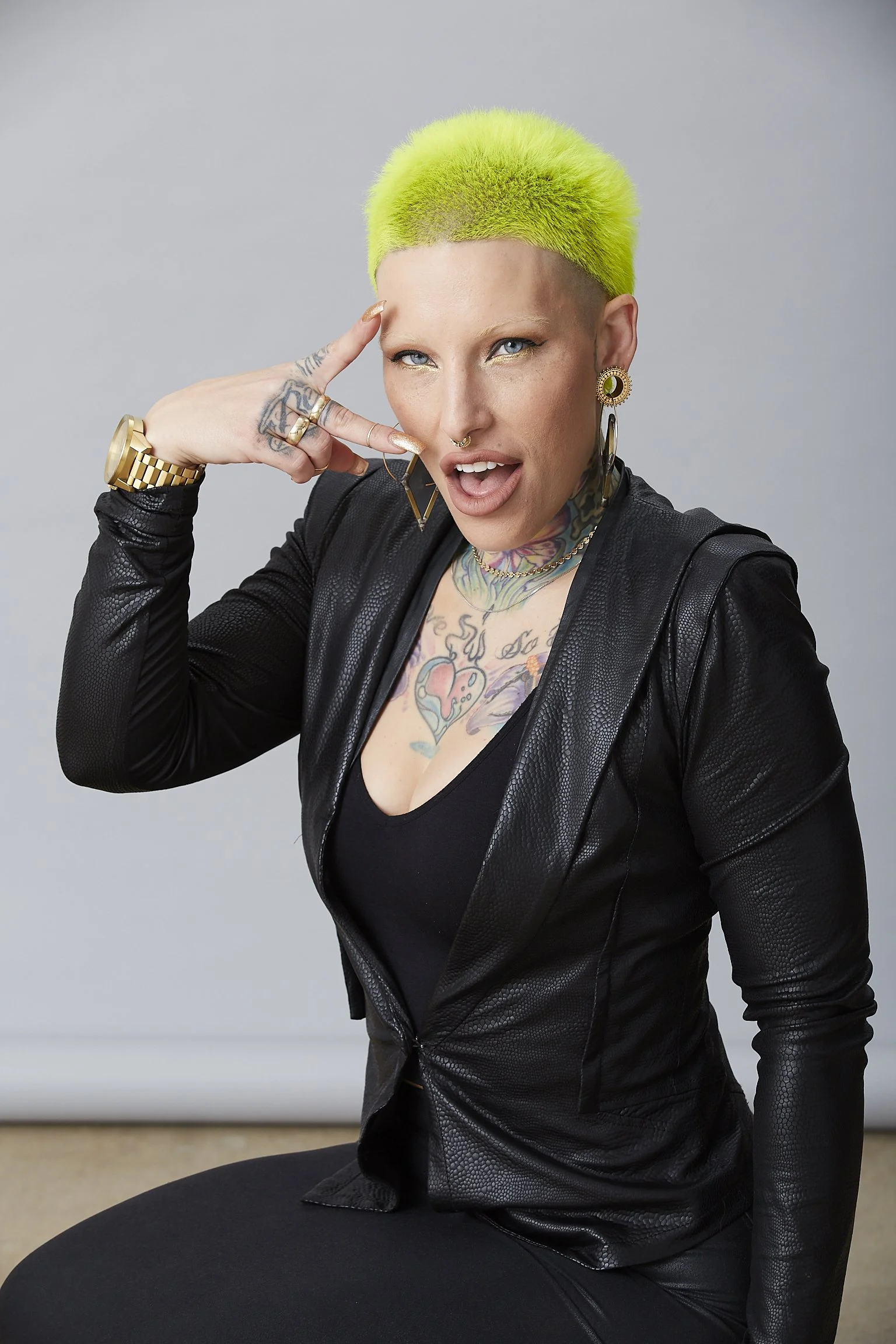 A woman with short, bright green hair styled in a buzz cut, wearing a black textured leather jacket and matching top, with tattoos on her chest and neck, jewelry including large earrings, rings, and a watch, making a playful gesture with her fingers 