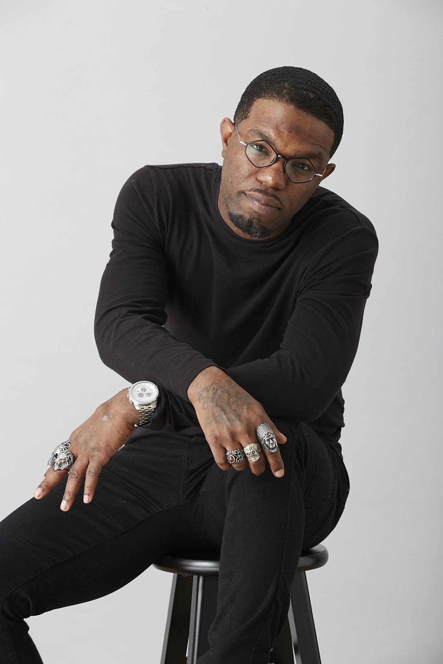 A man with dark skin, short black hair, glasses, wearing a black long-sleeve shirt and black pants, sitting on a stool against a plain light gray background, has tattoos on his left hand and wears multiple rings and a watch.