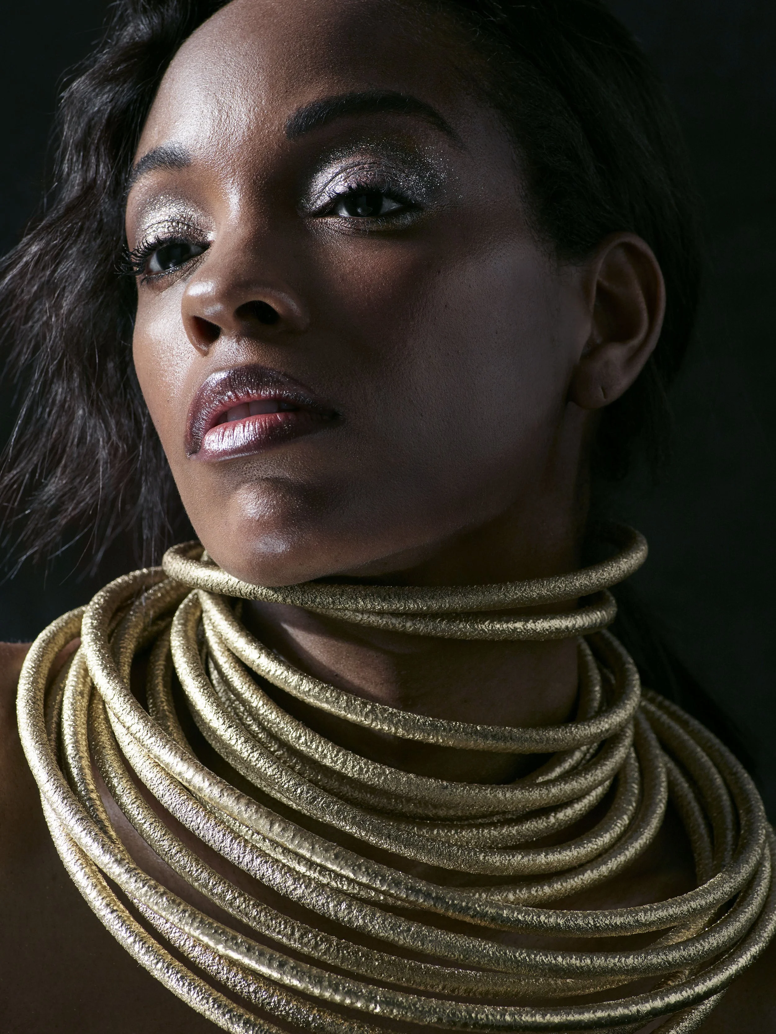 A close-up portrait of a woman with dark skin, wearing multiple gold metallic necklaces, with silver eyeshadow, dark lipstick, and textured hair.