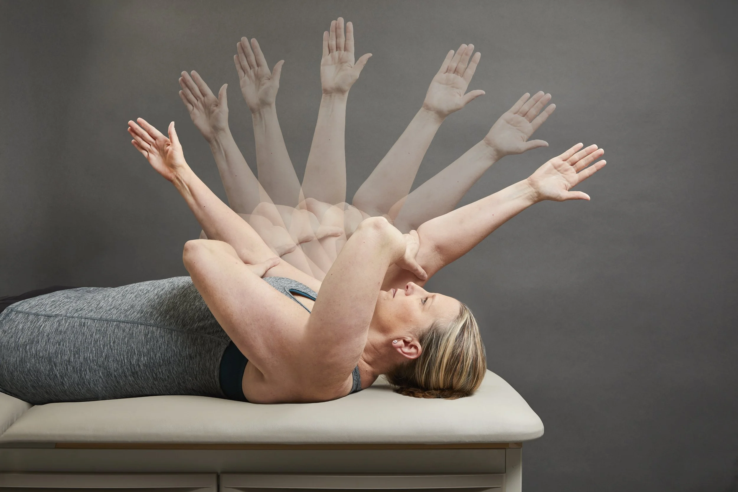 A woman lying on her back on a bed with her arms raised above her head, hands open, performing a stretching or exercise move. The background is a plain gray wall. Orthopedic. Orthopedic therapy. Shoulder injury