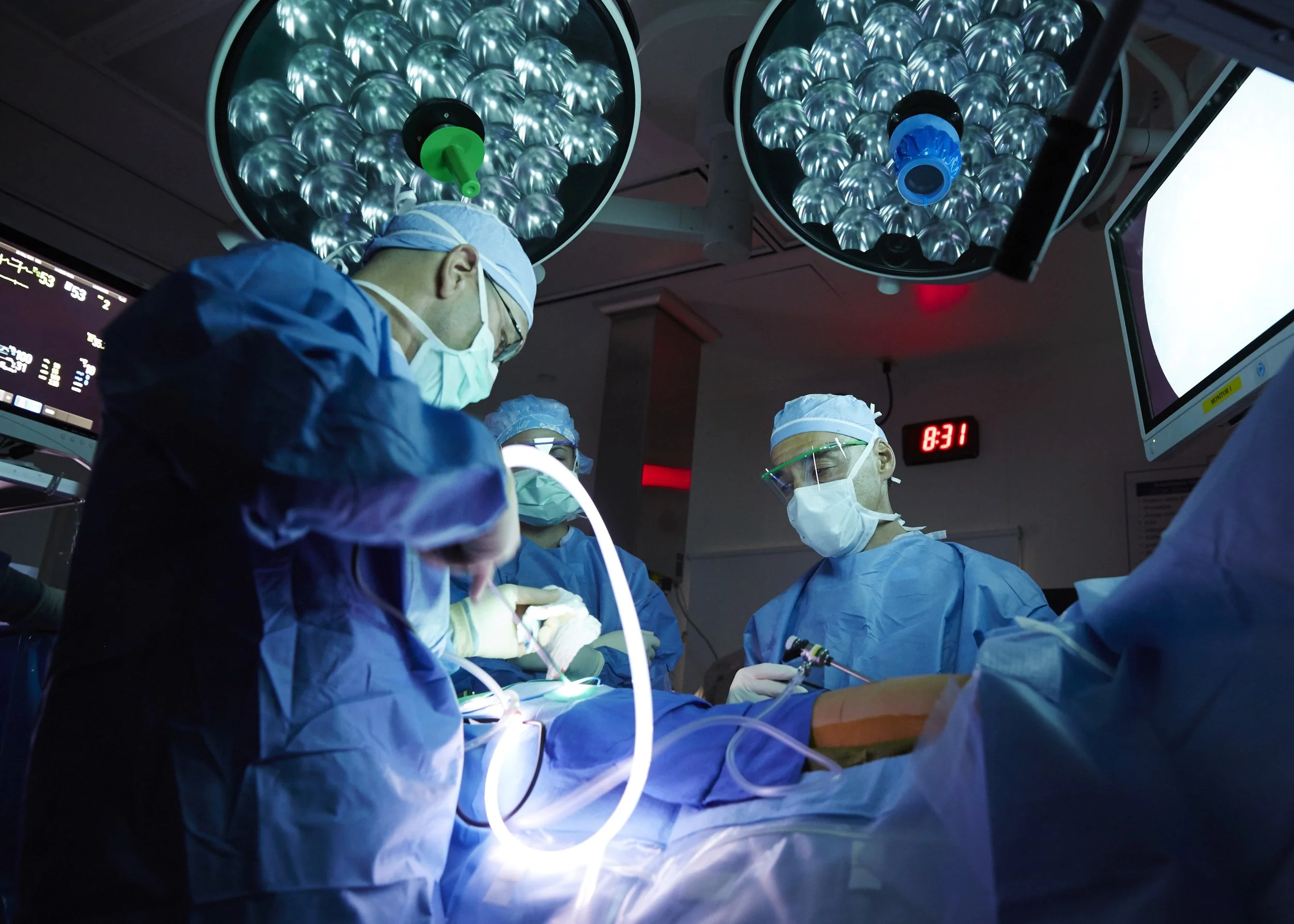 Medical team performing surgery in an operating room with bright overhead lights, monitors, and medical equipment.