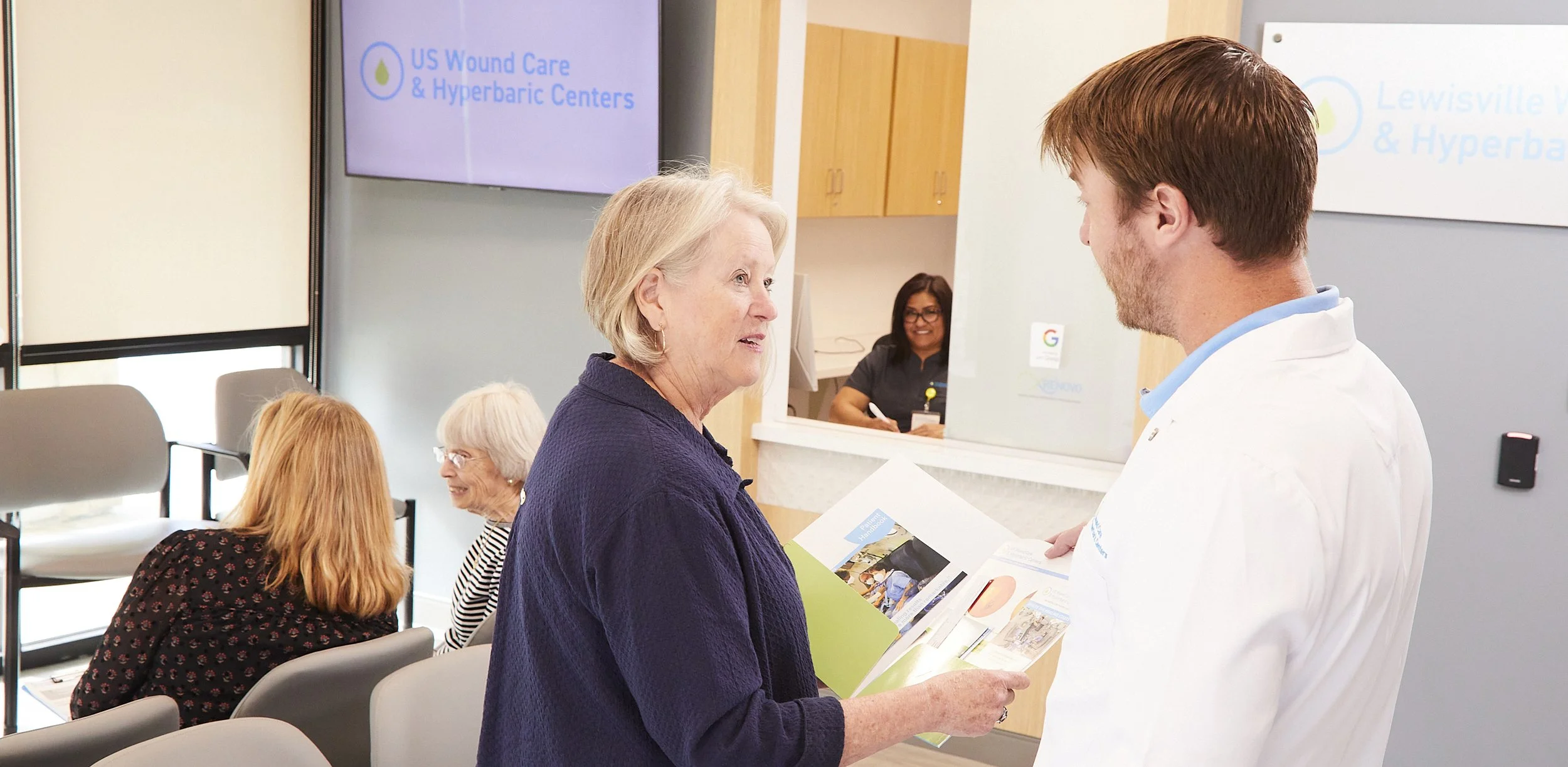 A healthcare professional in a white coat assisting an elderly woman at a medical center reception area. Two other women are seated nearby, and a receptionist is visible in the background. A sign on the wall reads 'US Wound Care & Hyperbaric Centers.