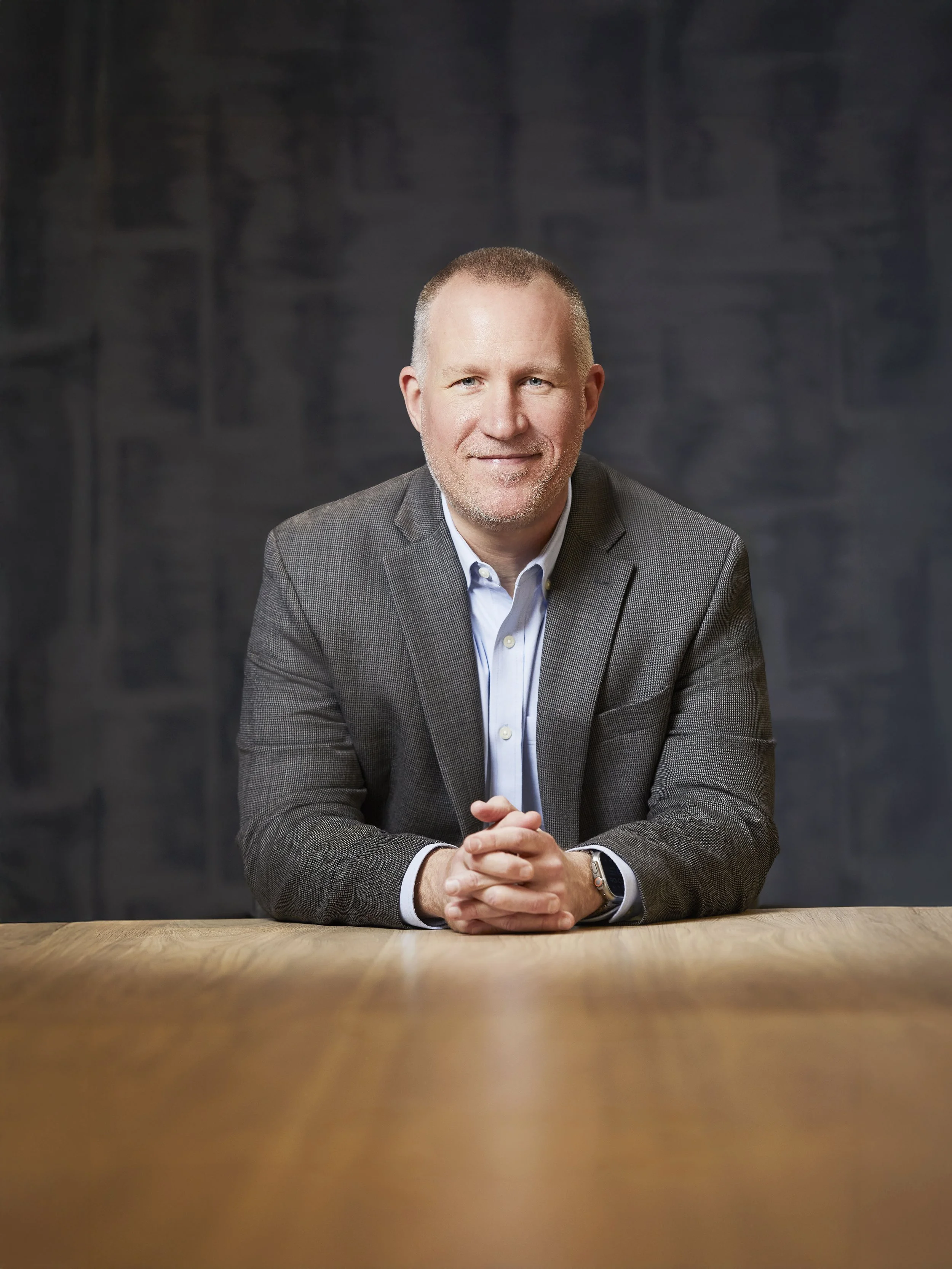A middle-aged man with a light beard and short hair, wearing a gray blazer over a light blue shirt, sitting at a wooden table with his hands clasped, smiling at the camera against a dark background.