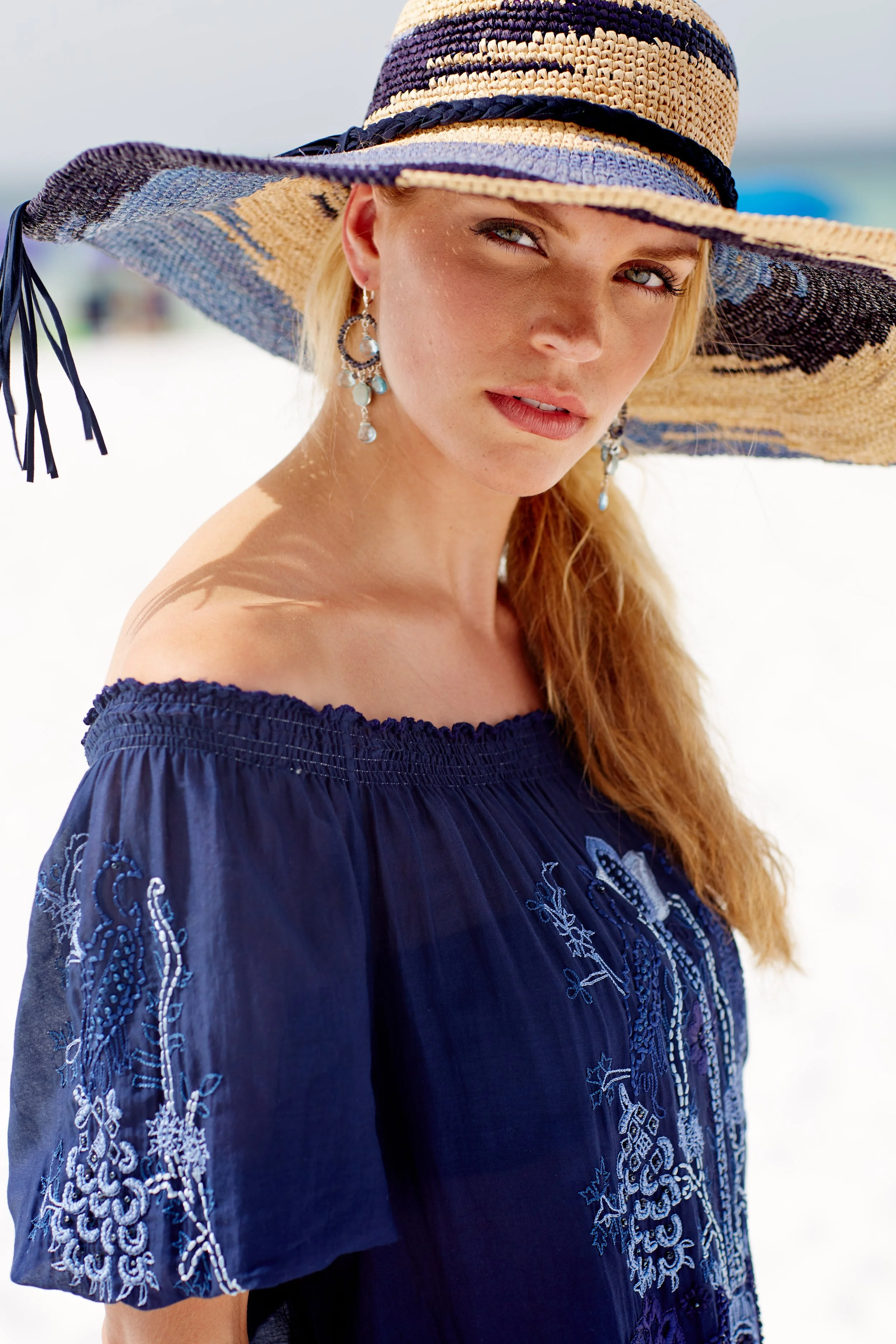 A woman wearing a wide-brimmed straw hat, large earrings, and a navy off-shoulder embroidered dress, standing in a bright outdoor setting with a blurry background.
