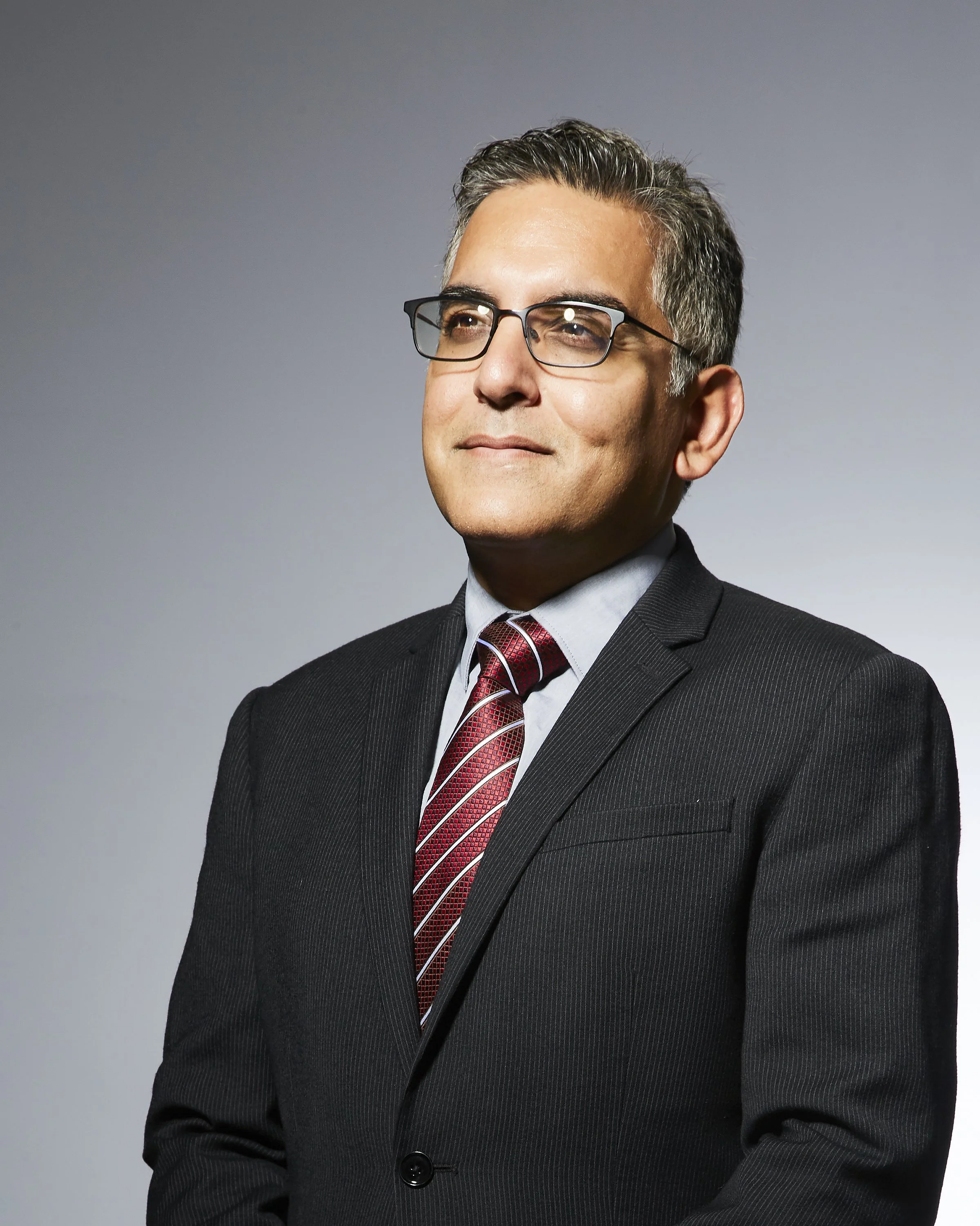 A professional man wearing glasses, a dark suit, a light blue shirt, and a red patterned tie, looking thoughtfully to the side against a neutral gray background.