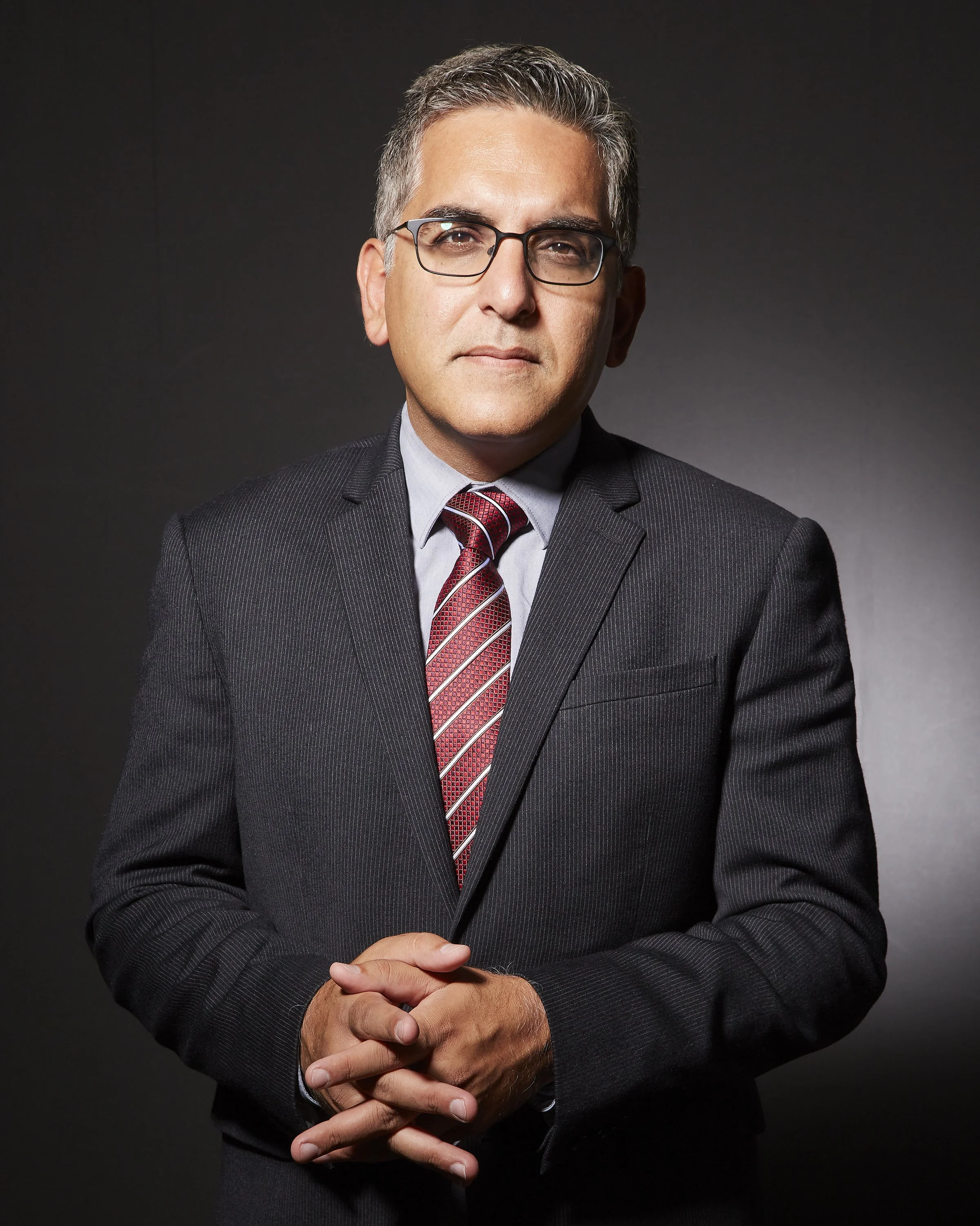 A man with glasses in a dark suit and a red patterned tie, standing against a dark background.
