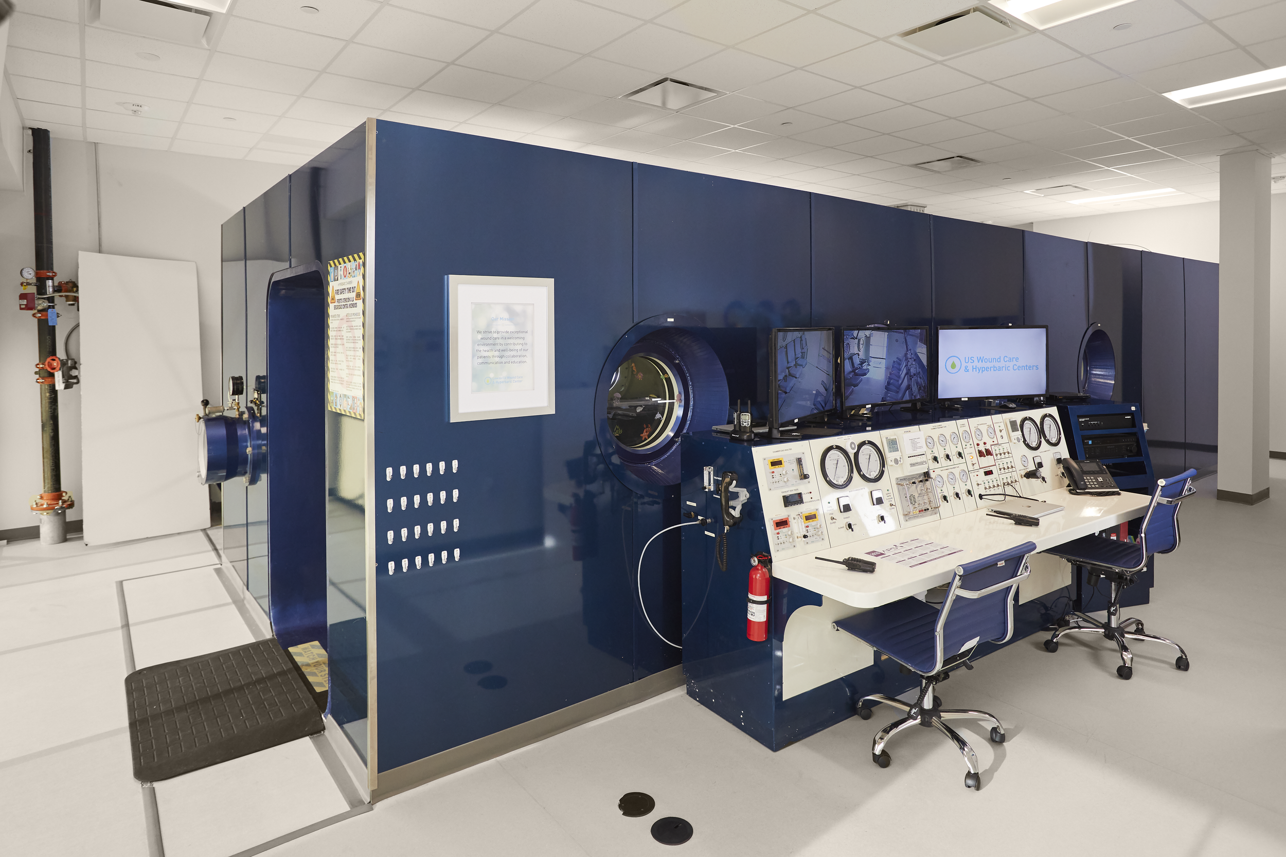 Medical testing chamber with control panel, monitors, and seating. The chamber has a circular glass window, and the control panel includes dials, buttons, and communication devices. A fire extinguisher is attached to the side of the control panel.
