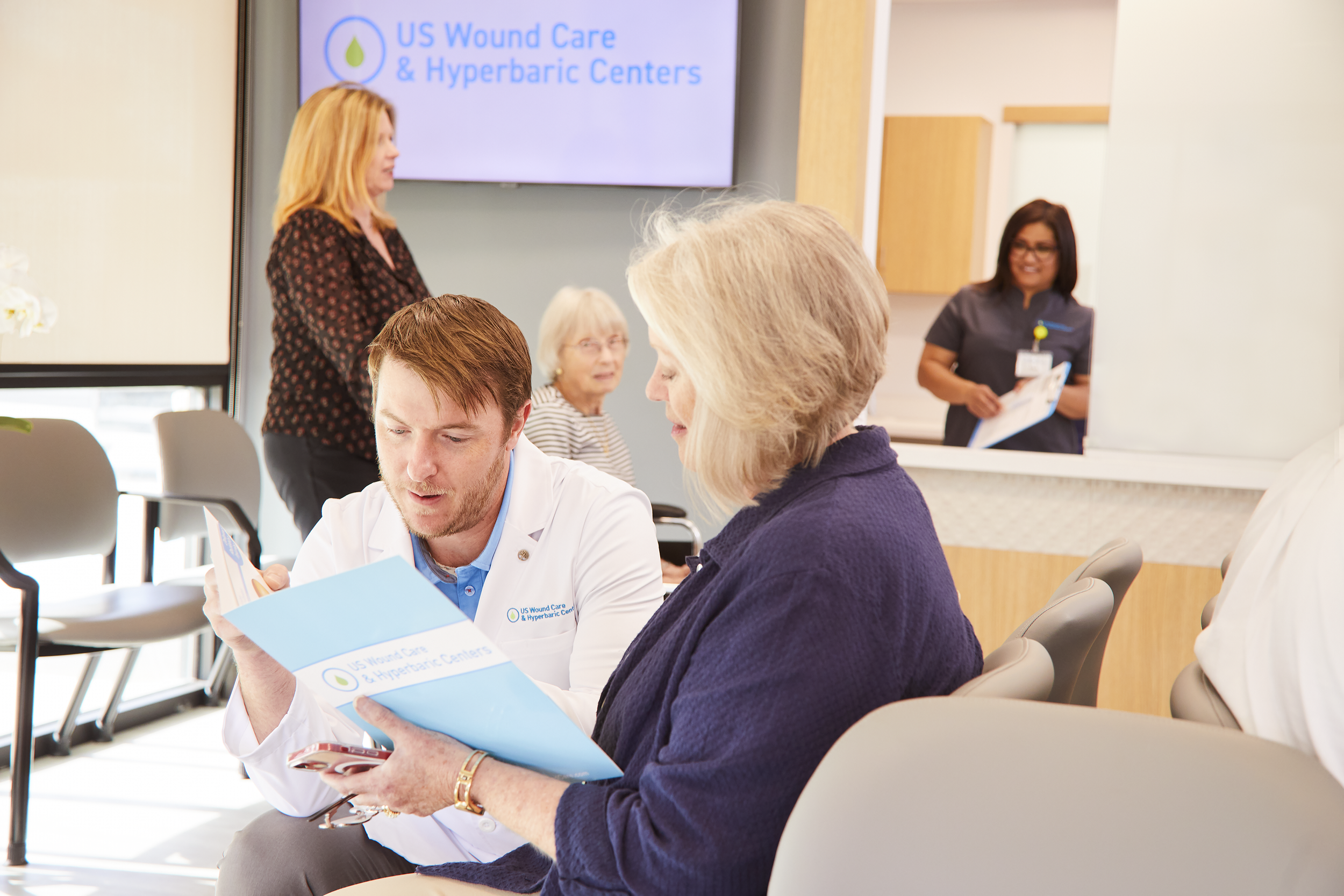 Healthcare professionals and patients in a medical waiting area of US Wound Care & Hyperbaric Centers, with a large digital screen displaying the center's name and logo.