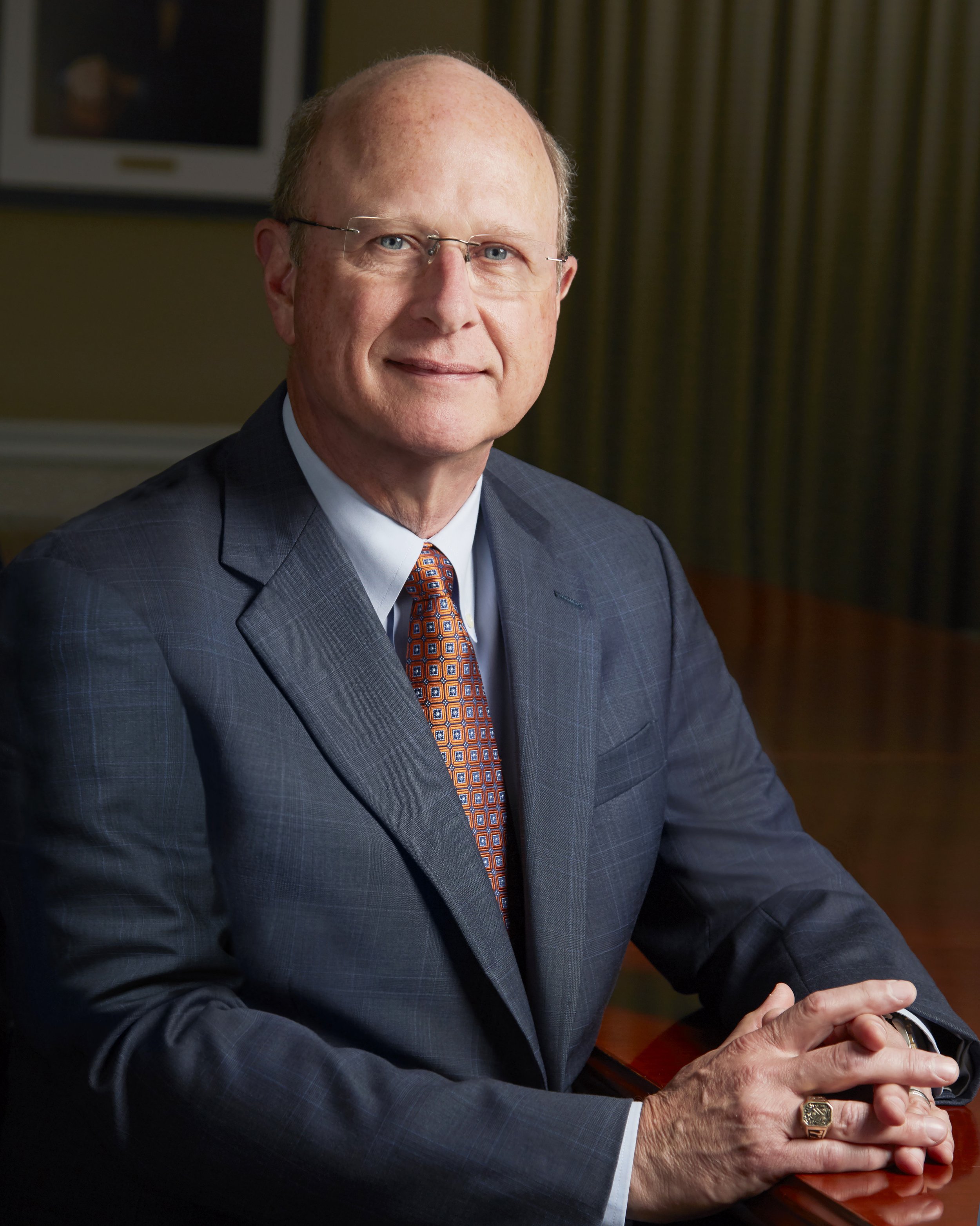 A middle-aged man with glasses wearing a dark blue suit, white shirt, and orange patterned tie, seated at a wooden table in a formal setting with curtains in the background.