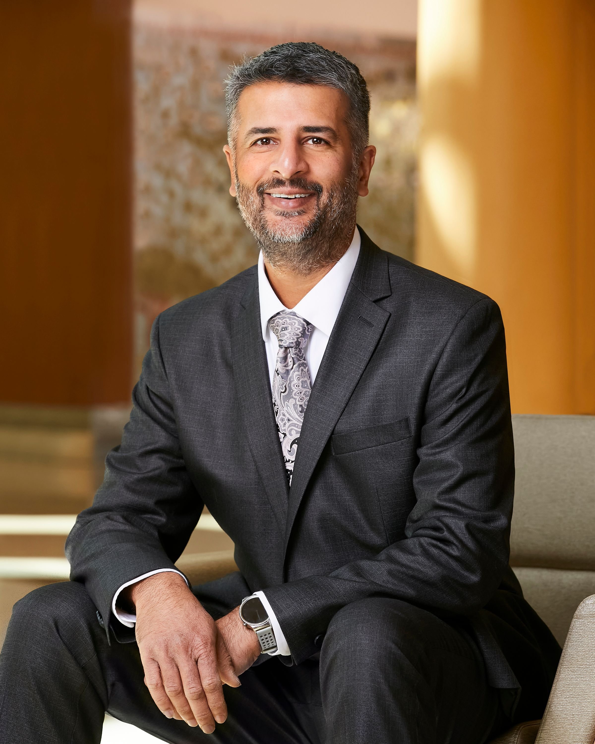 A man with gray hair and a beard wearing a dark suit, white shirt, and patterned tie, sitting on a beige chair in a professional setting, smiling at the camera.