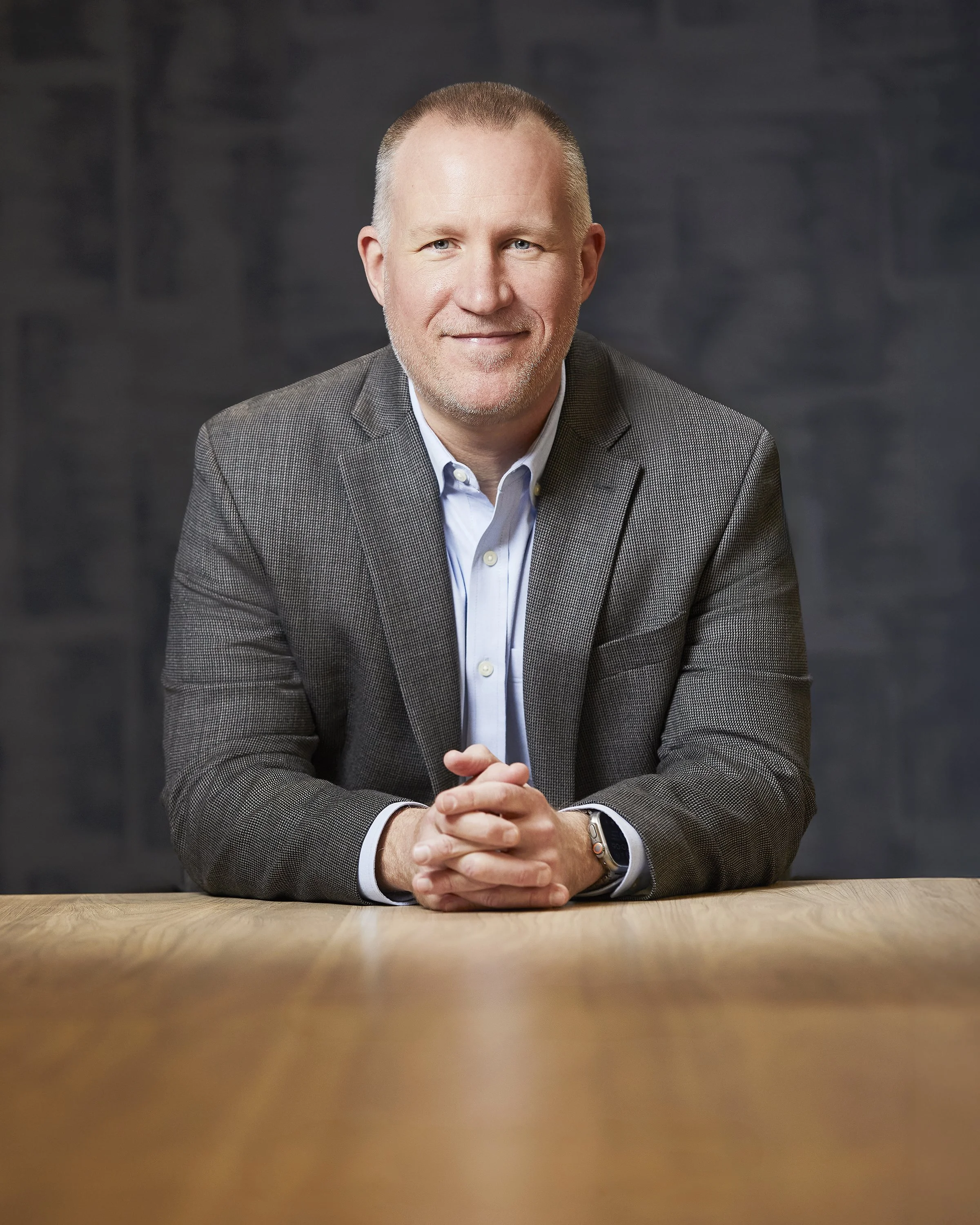 Portrait of a middle-aged man in a gray suit jacket and light blue shirt sitting at a wooden table, smiling softly, with a dark blurred background.