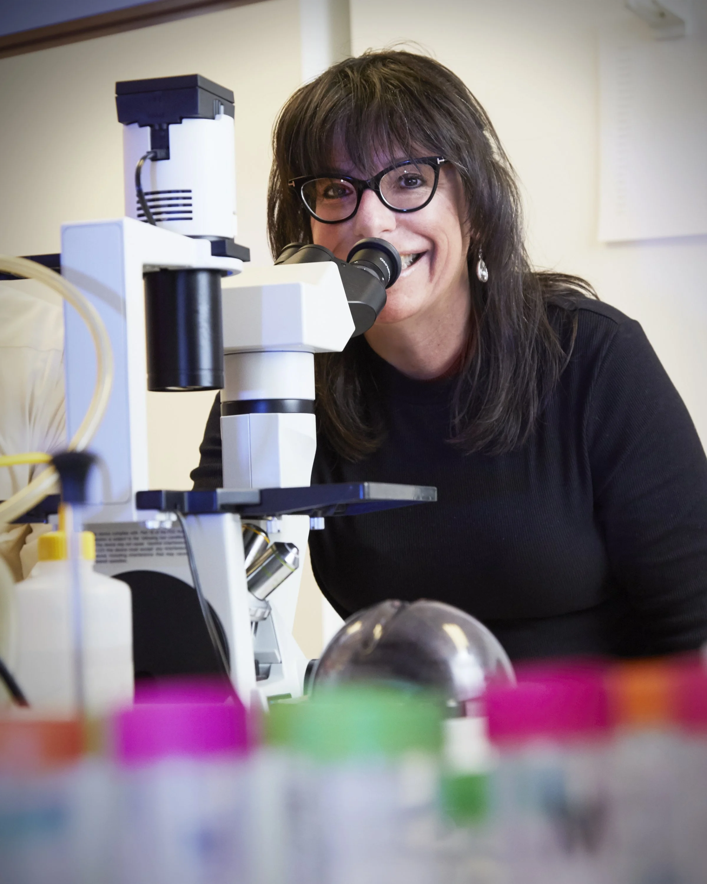 A woman with glasses and dark hair looking through a microscope in a laboratory.