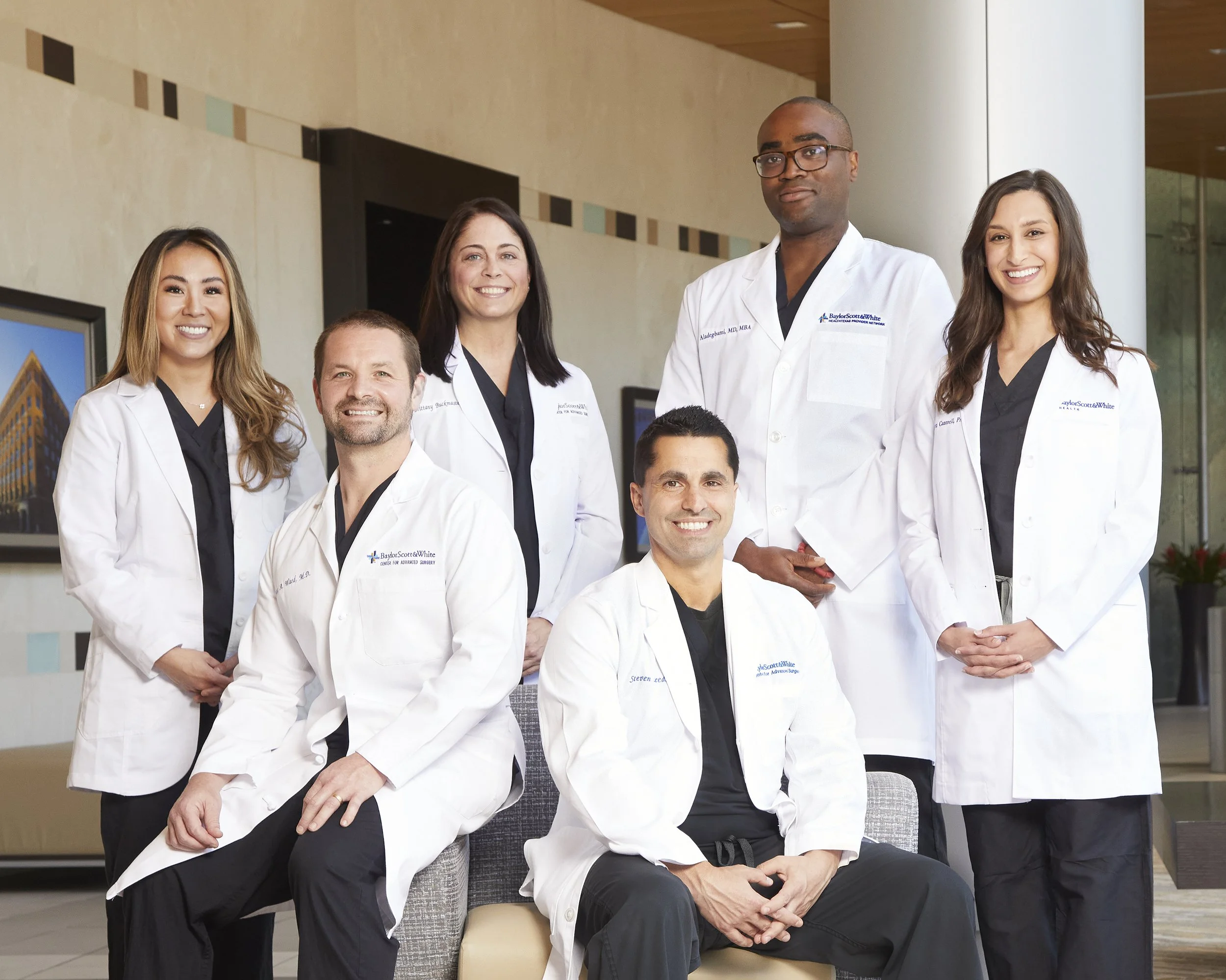 Group of seven diverse medical professionals dressed in white coats in a modern hospital lobby, smiling for a photo.