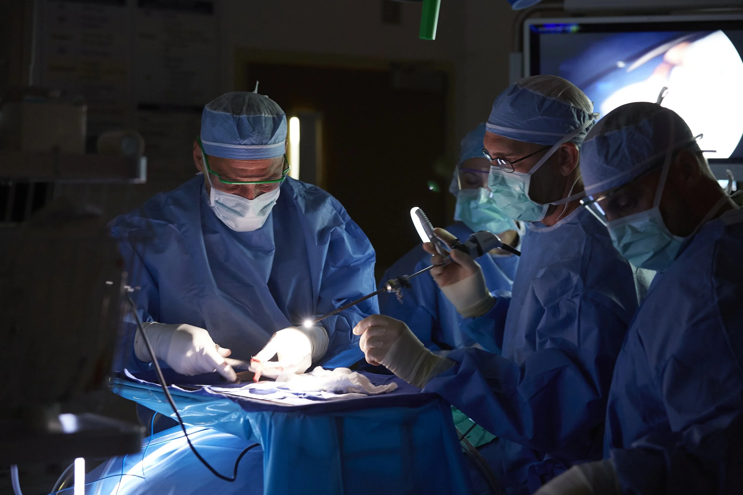 A team of surgeons performing an operation in an operating room, with one surgeon holding surgical tools and others assisting, all dressed in blue surgical gowns, masks, and caps.