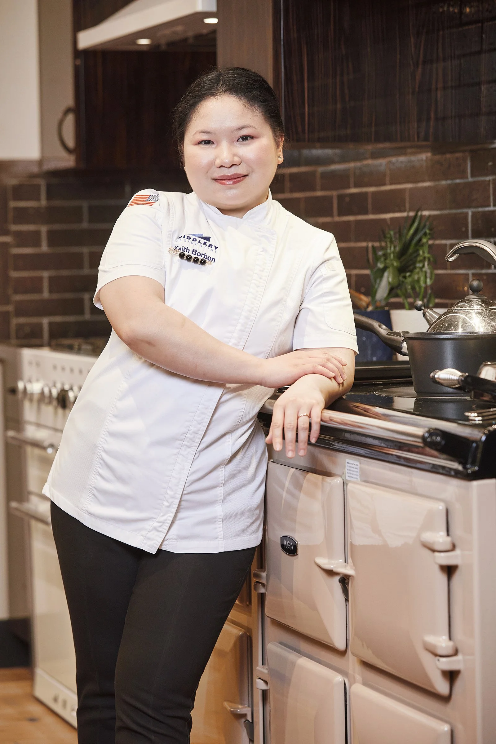 A woman in a white chef's jacket standing in a kitchen, leaning on a vintage pastel-colored stove with pots and plants in the background.