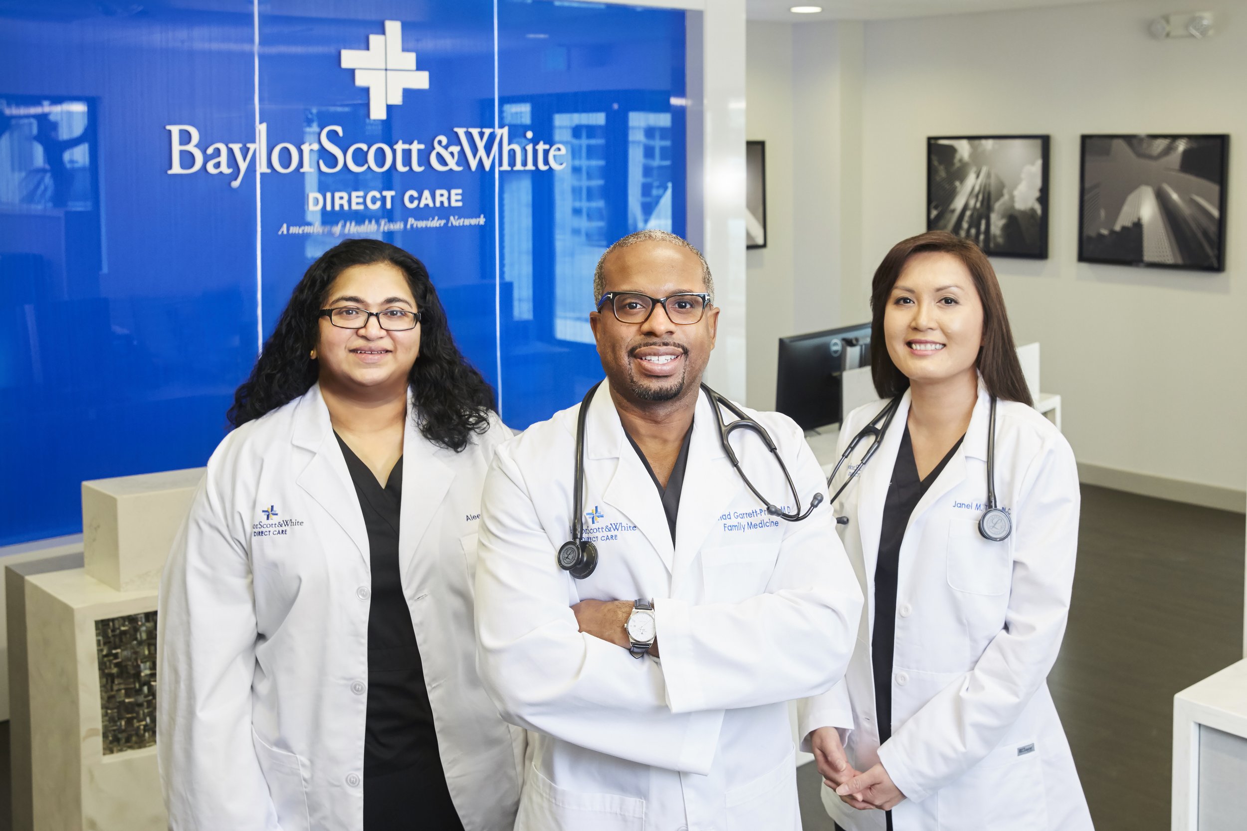 Three doctors standing inside Baylor Scott & White healthcare facility, wearing white coats and stethoscopes, smiling at the camera.