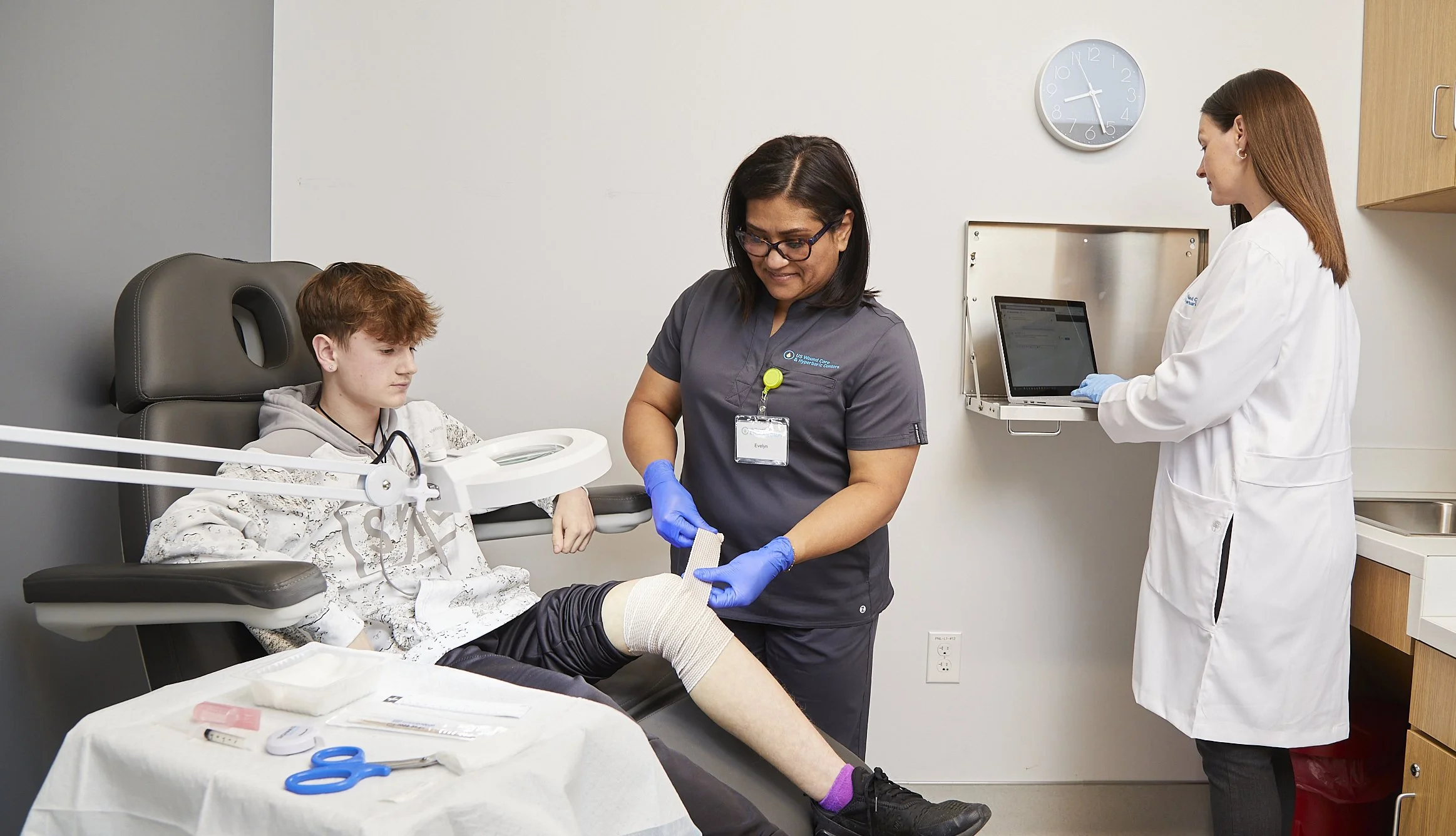 A young male patient sitting in an examination chair with his leg elevated and wrapped with a bandage, being examined by a nurse. A healthcare professional in a white coat is working on a laptop in the background, in a medical clinic or hospital room