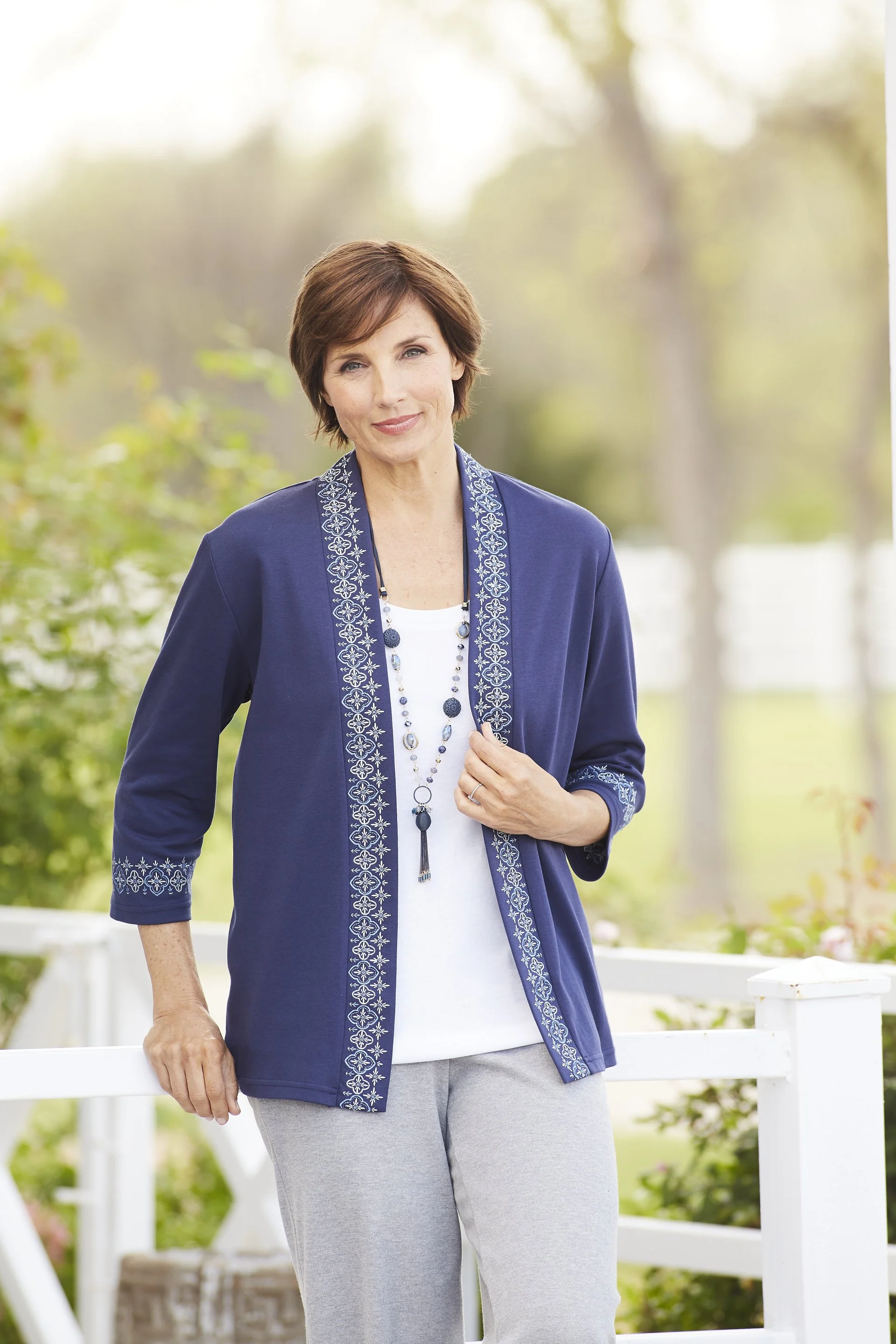 A woman standing outdoors on a porch, wearing a navy cardigan with embroidery, a white shirt, gray pants, and a long necklace, with greenery in the background.