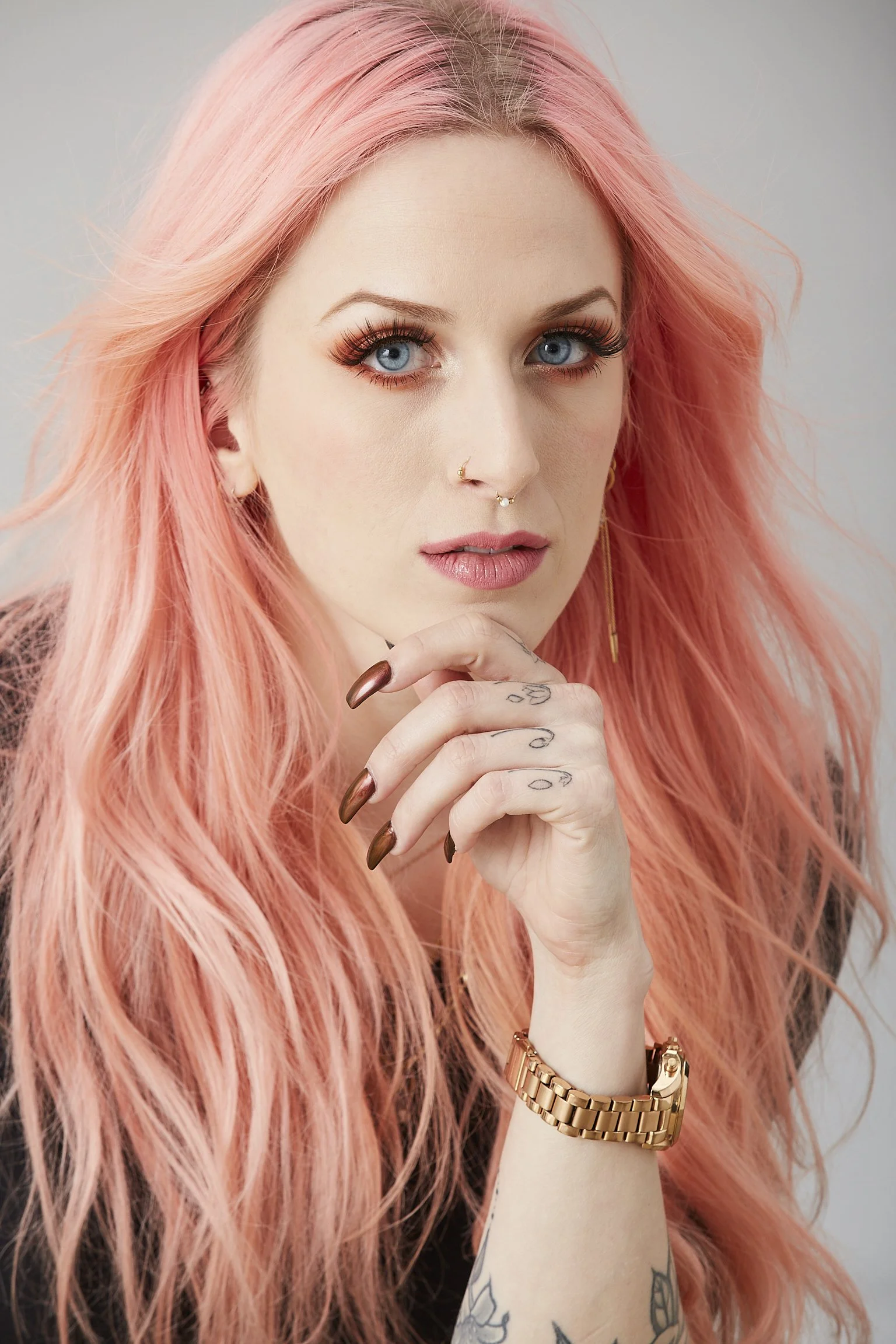 A woman with long pink hair, blue eyes, and light makeup, wearing a black top, gold watch, and earrings, with tattoos on her hand, looking intently at the camera with her hand near her chin.