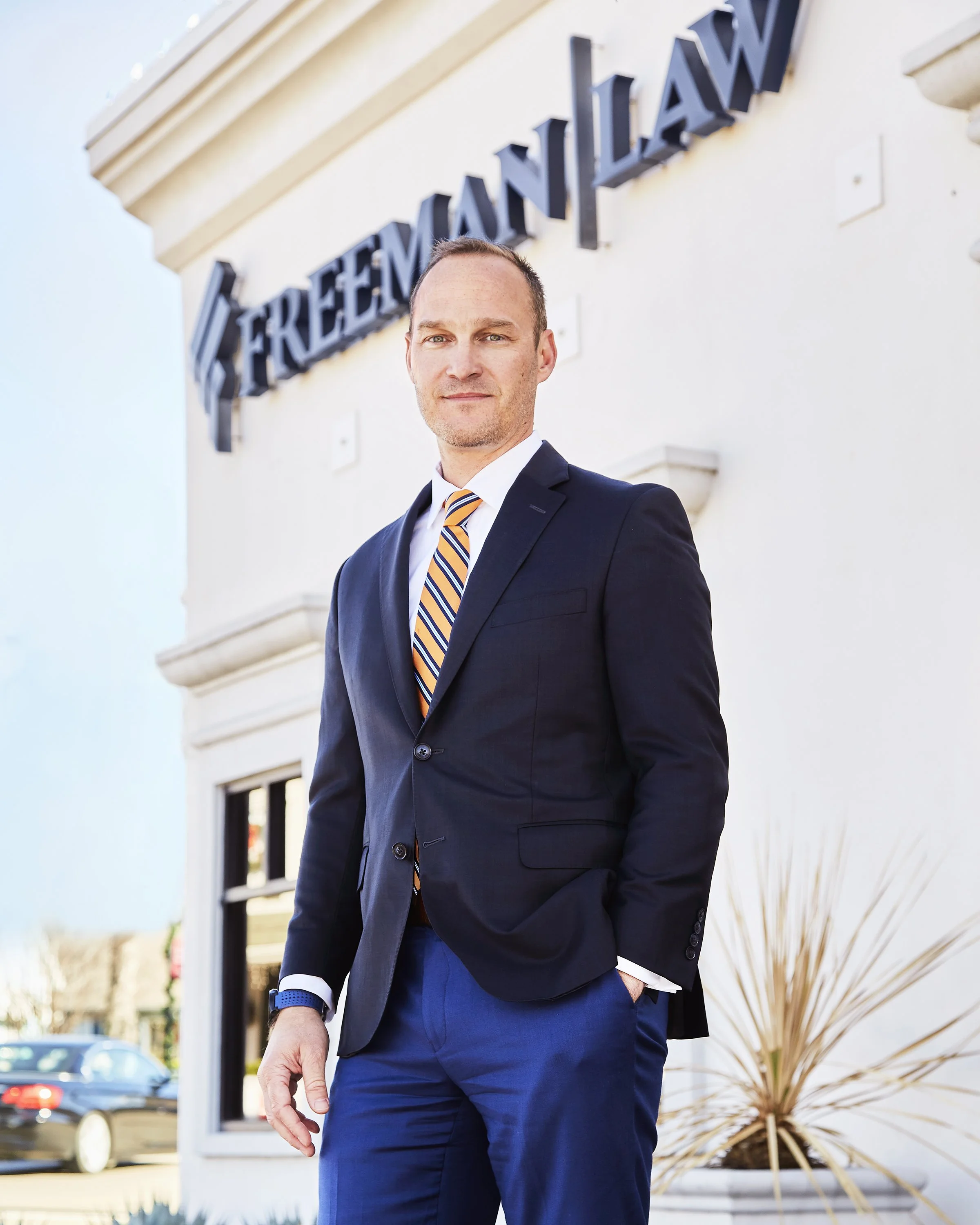 A man in a dark suit with a striped tie stands outside of a building with a sign that says 'FREEMAN LAW'.