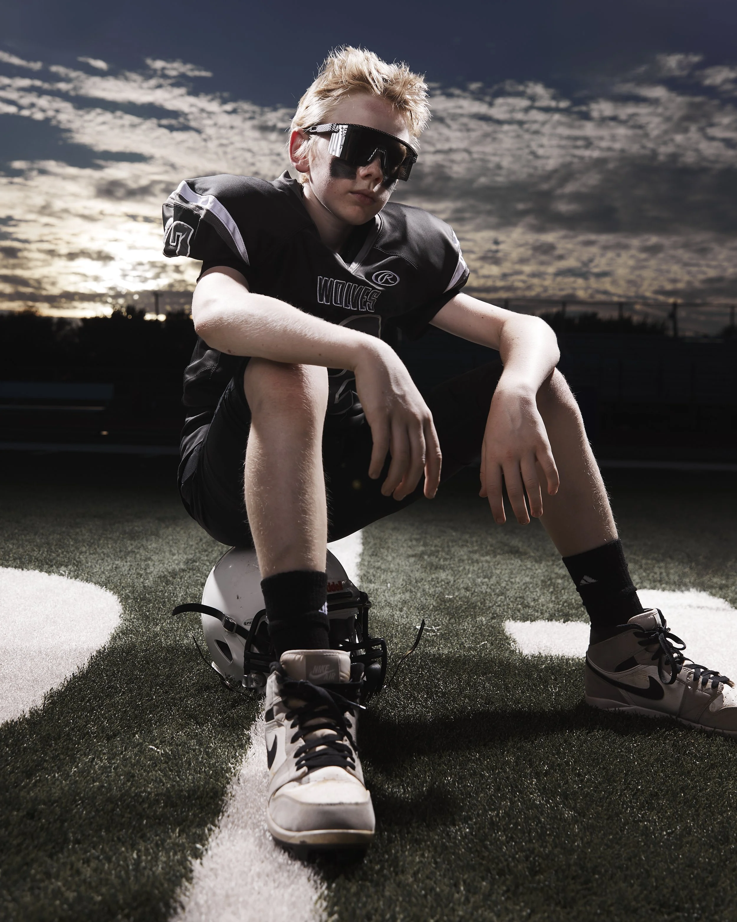 A young male football player in black uniform, black sunglasses, and white Nike shoes sitting on a football helmet on a field at sunset.